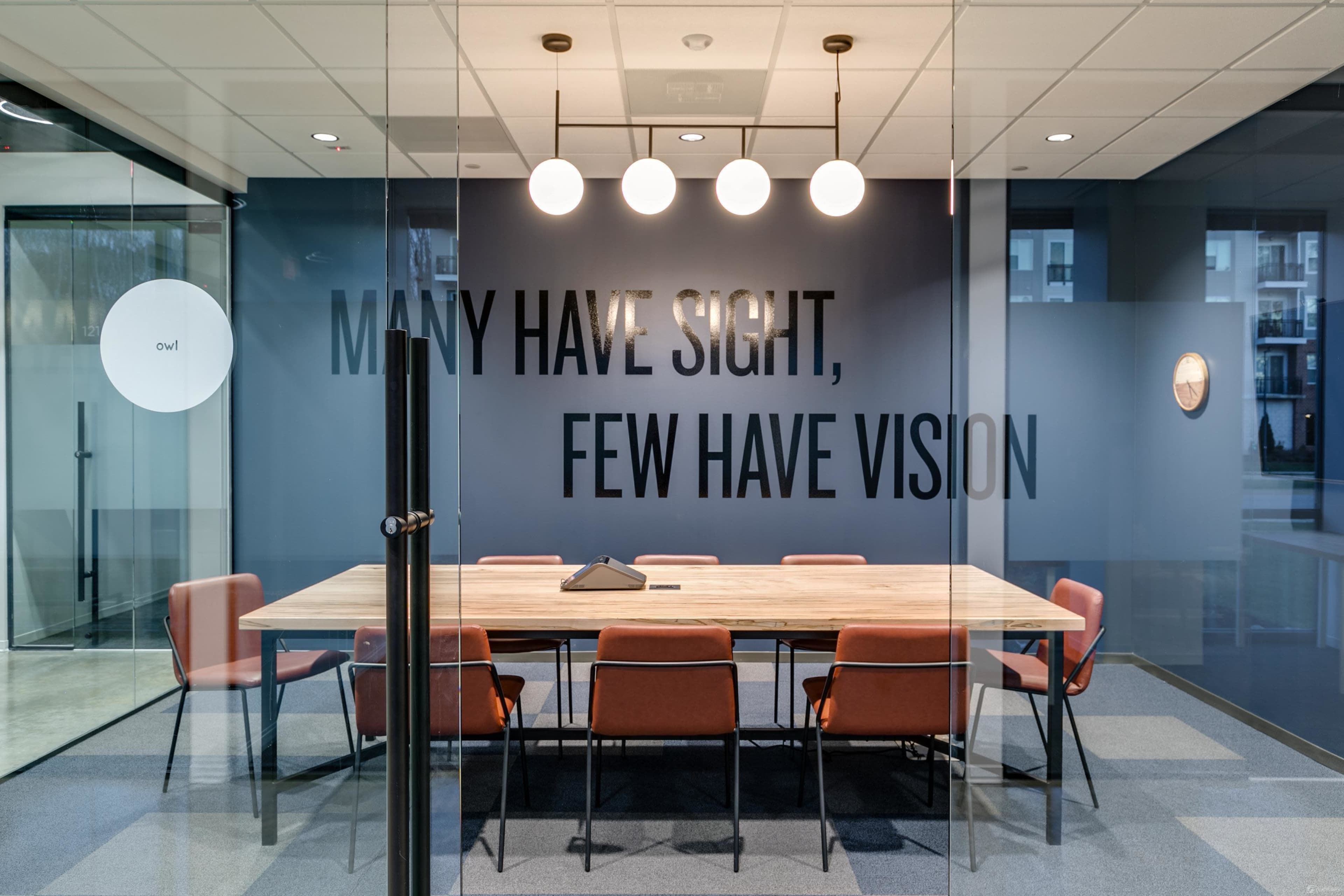 A modern conference room features a large wooden table surrounded by orange chairs, with a wall displaying the words "MANY HAVE SIGHT, FEW HAVE VISION."