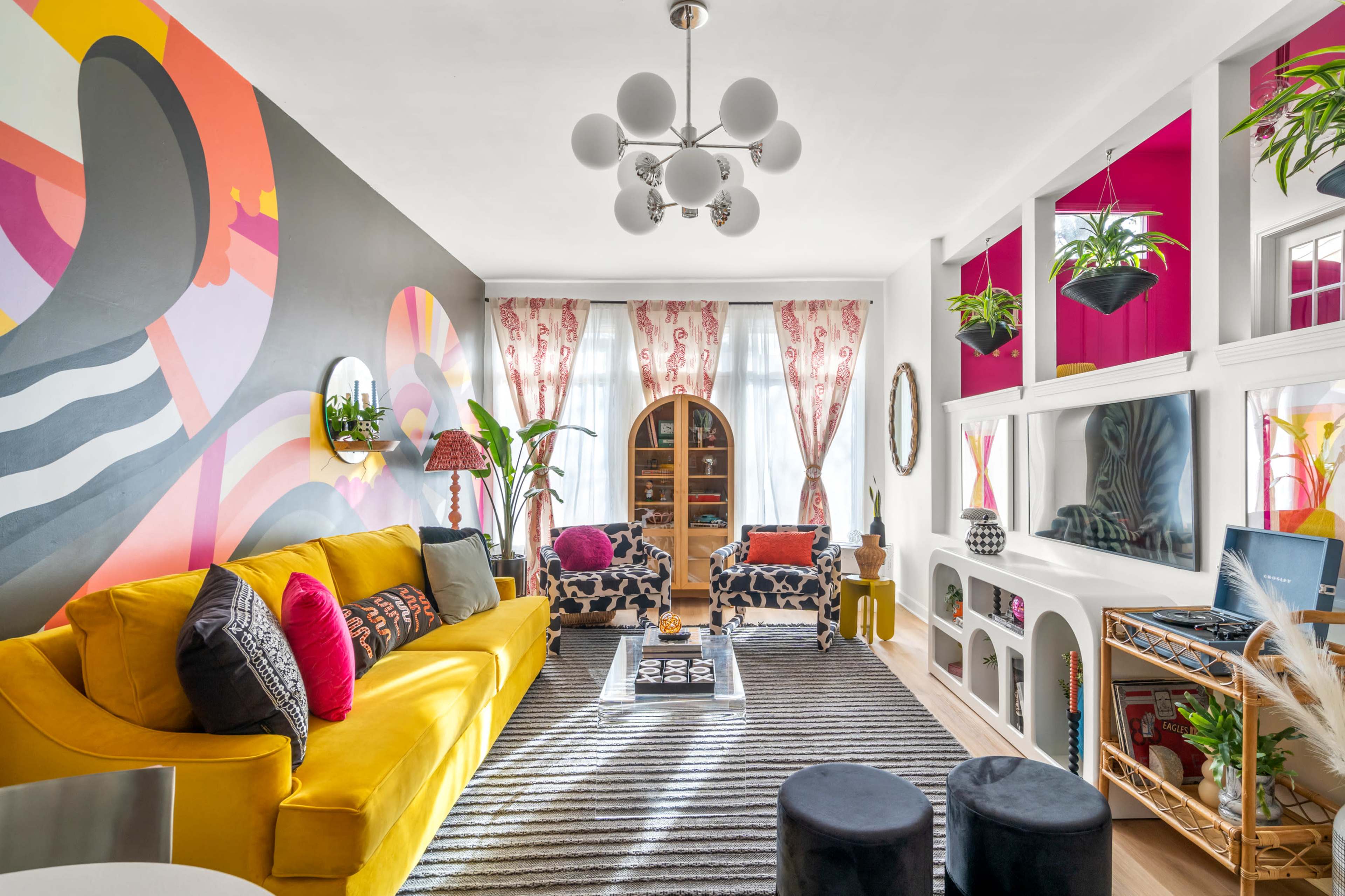 The living room features a bright yellow couch, patterned chairs, and a large mural on one wall, complemented by decorative plants and a striped rug.