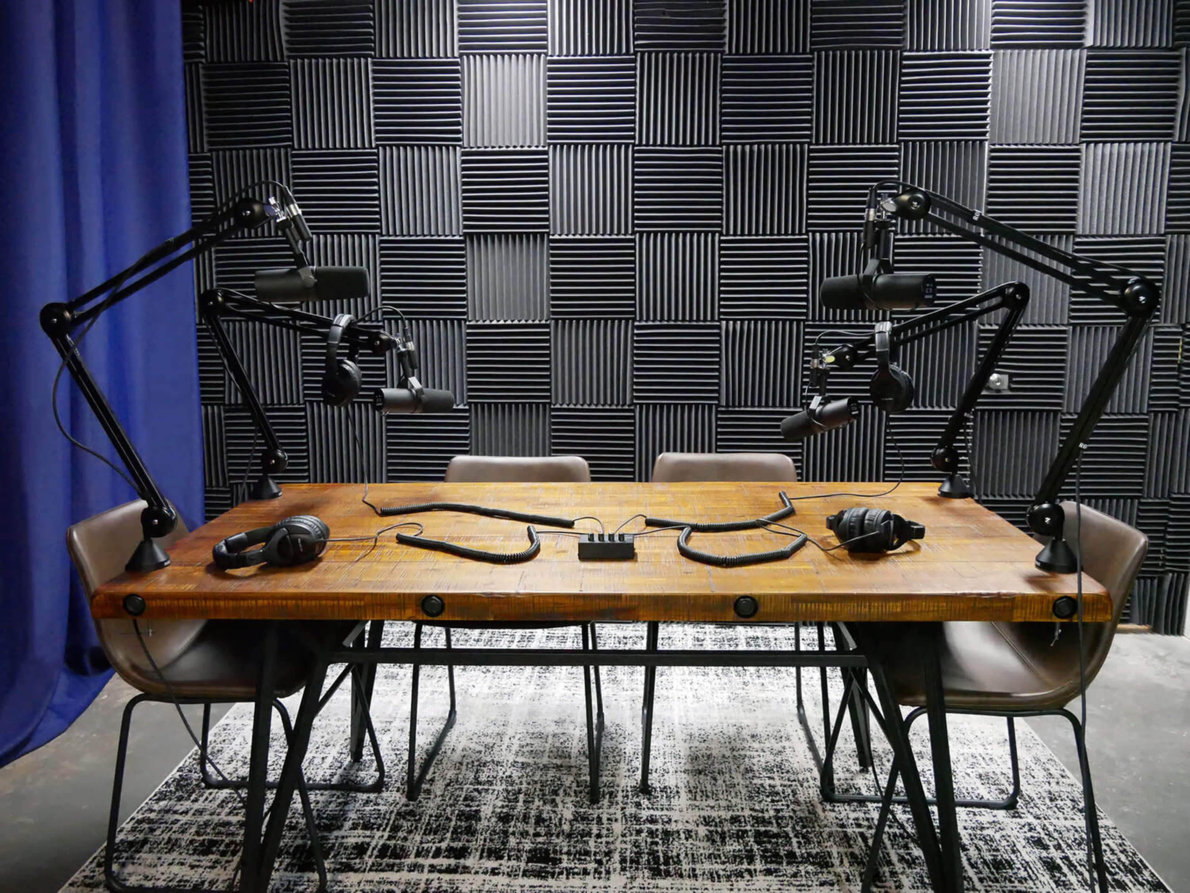 A spacious audio recording studio features a wooden table with microphones and headphones set up at each of the four chairs, against a backdrop of soundproofing panels.