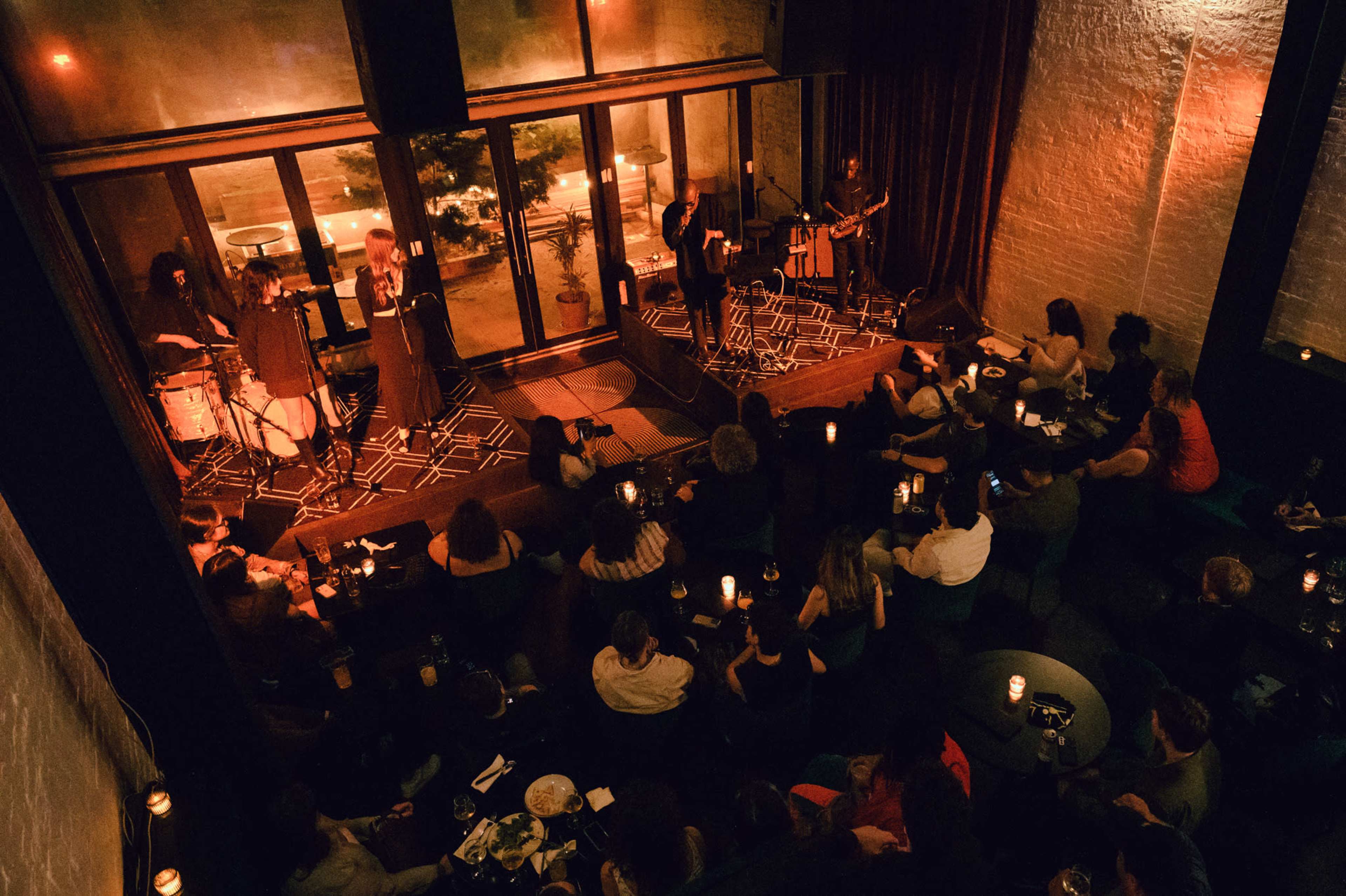 A band performs on a stage in a dimly lit venue while an audience listens from seated tables.