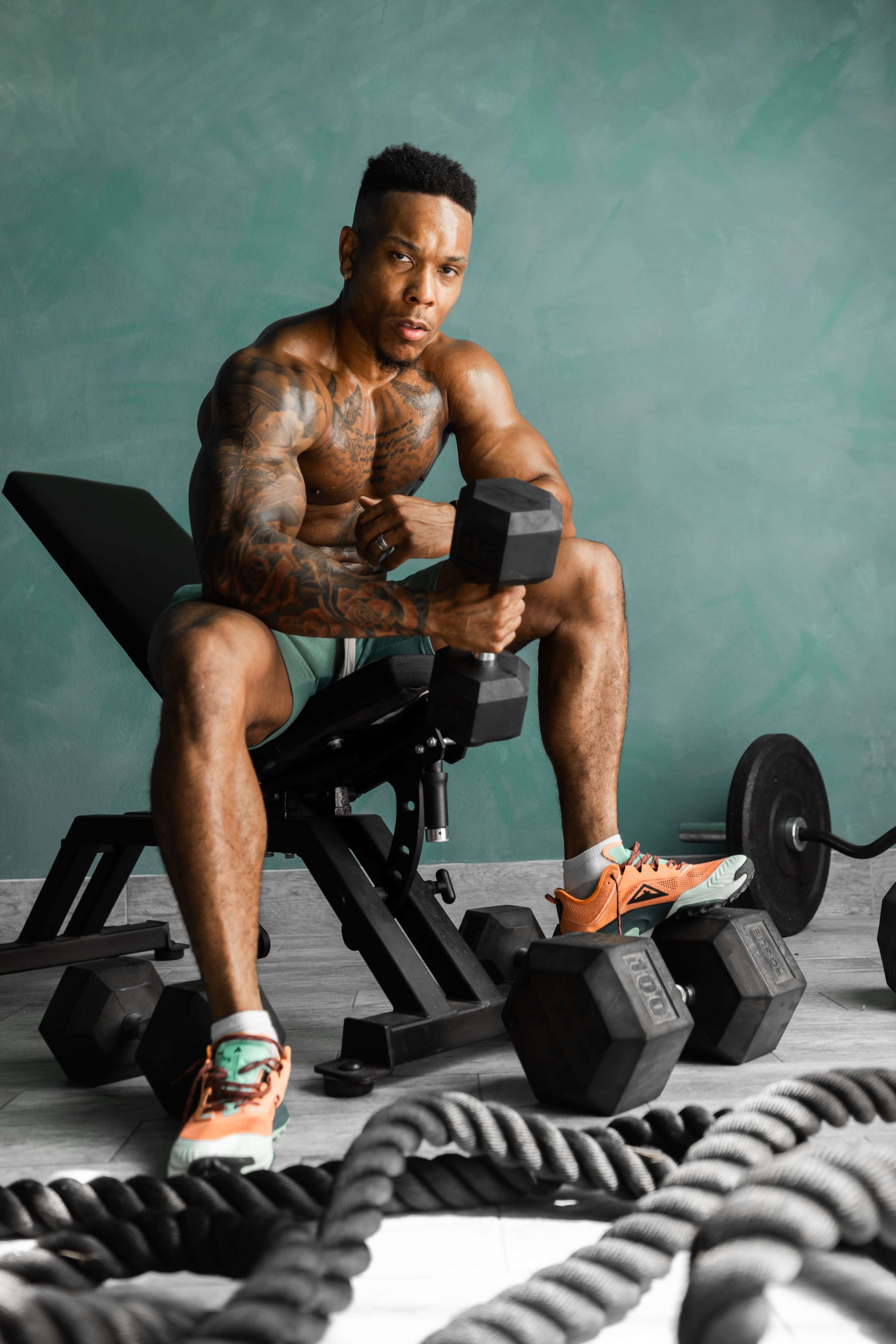 A muscular man with tattoos sits on a bench in a gym, holding a dumbbell among various weights and ropes.