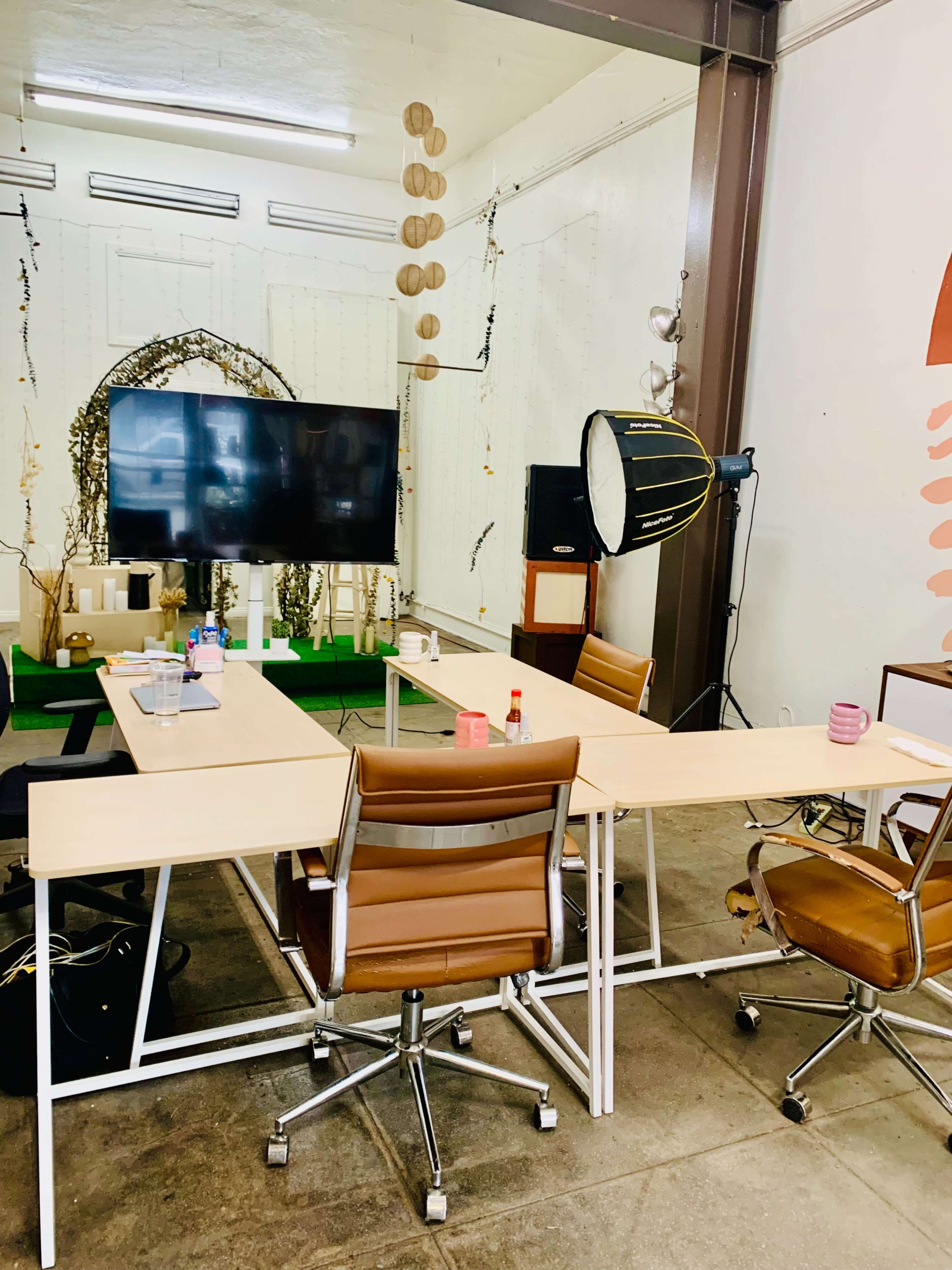 The image shows a minimalist meeting space with two large tables, brown leather chairs, a television screen, and a large studio light.