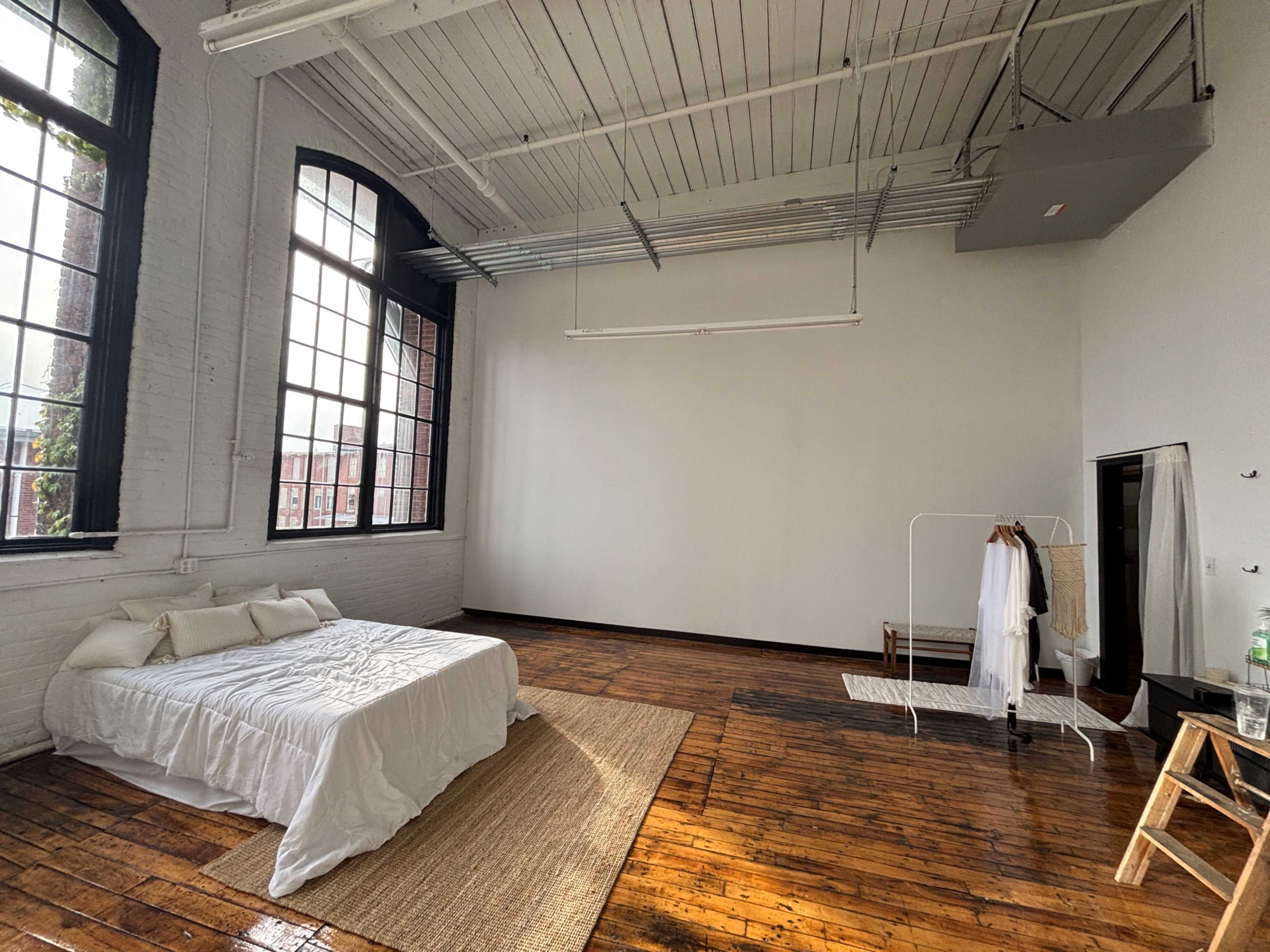 The image shows a spacious, well-lit bedroom with large windows, a white bedspread, wooden floors, and minimal decor.