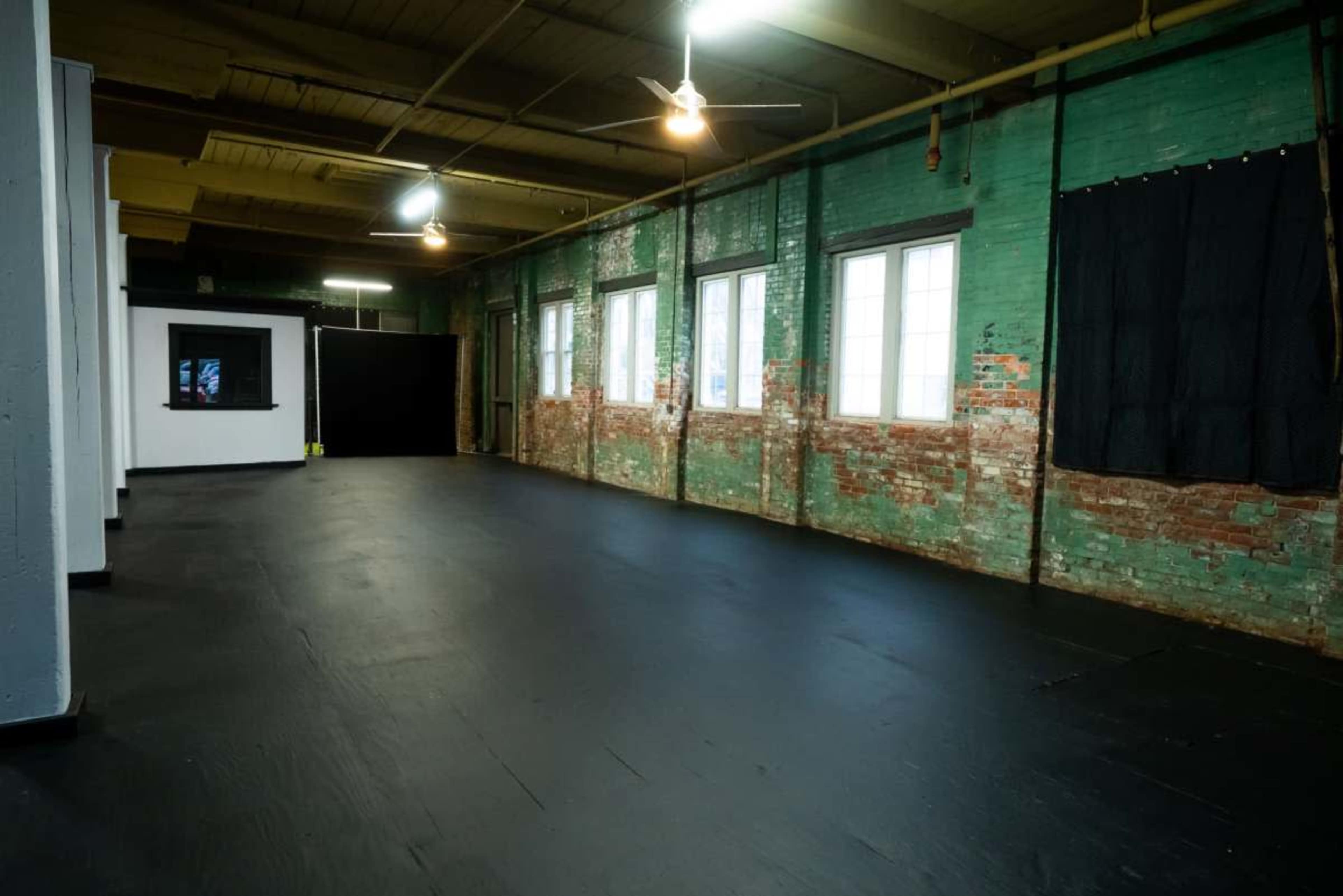 The image shows a spacious room with exposed brick walls, large windows, and a dark wooden floor.