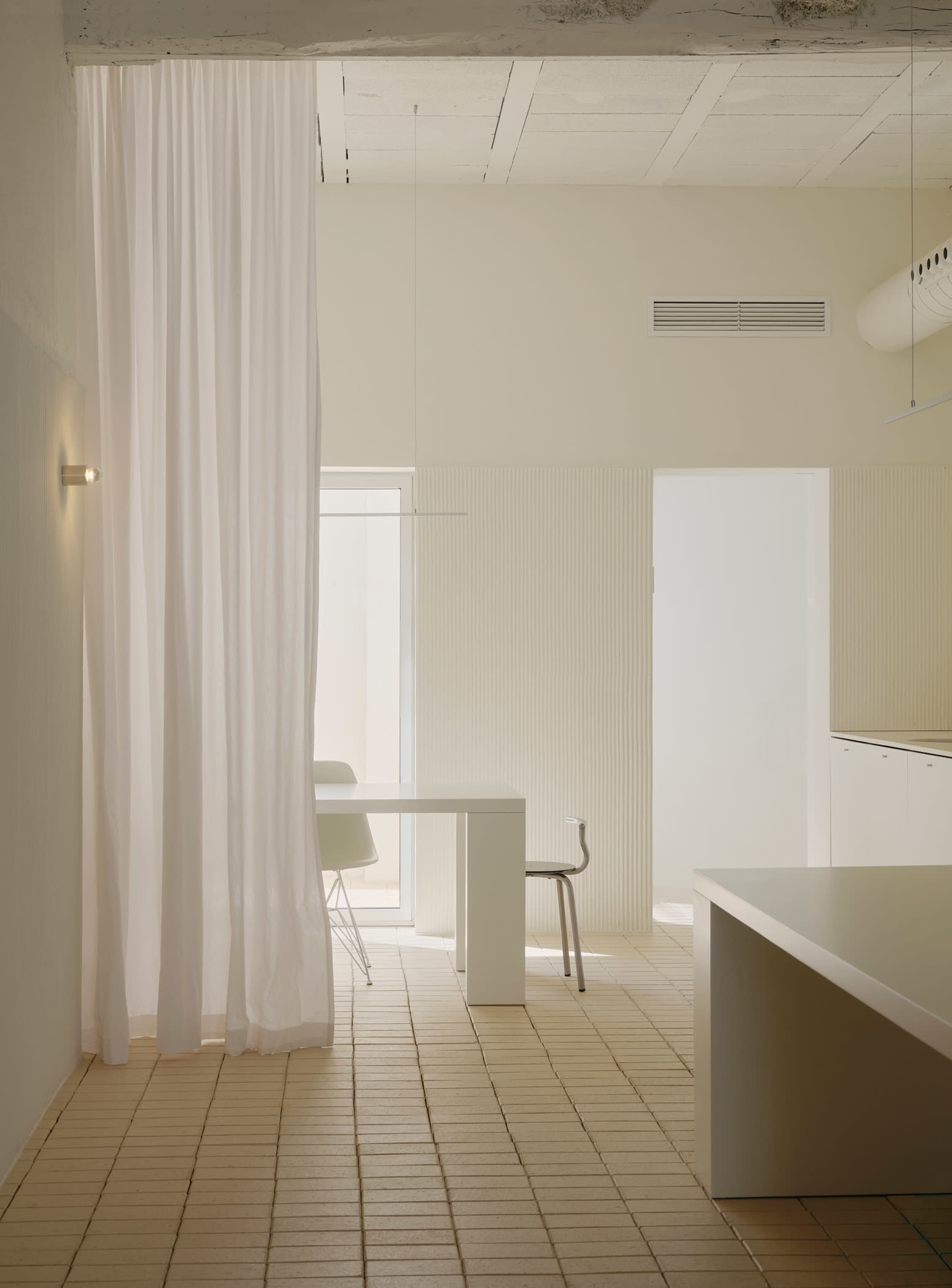 The image shows a minimalist interior space featuring a white table, a chair, and a large sheer curtain, with light-colored walls and tiled flooring.