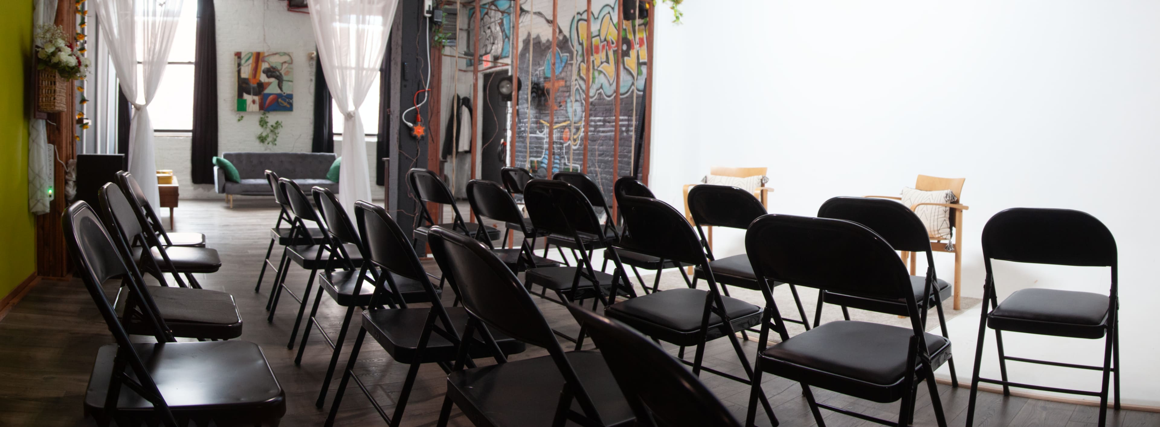 The image shows a room set up for an event with rows of black folding chairs facing a white wall, while a gray couch and colorful wall art are visible in the background.