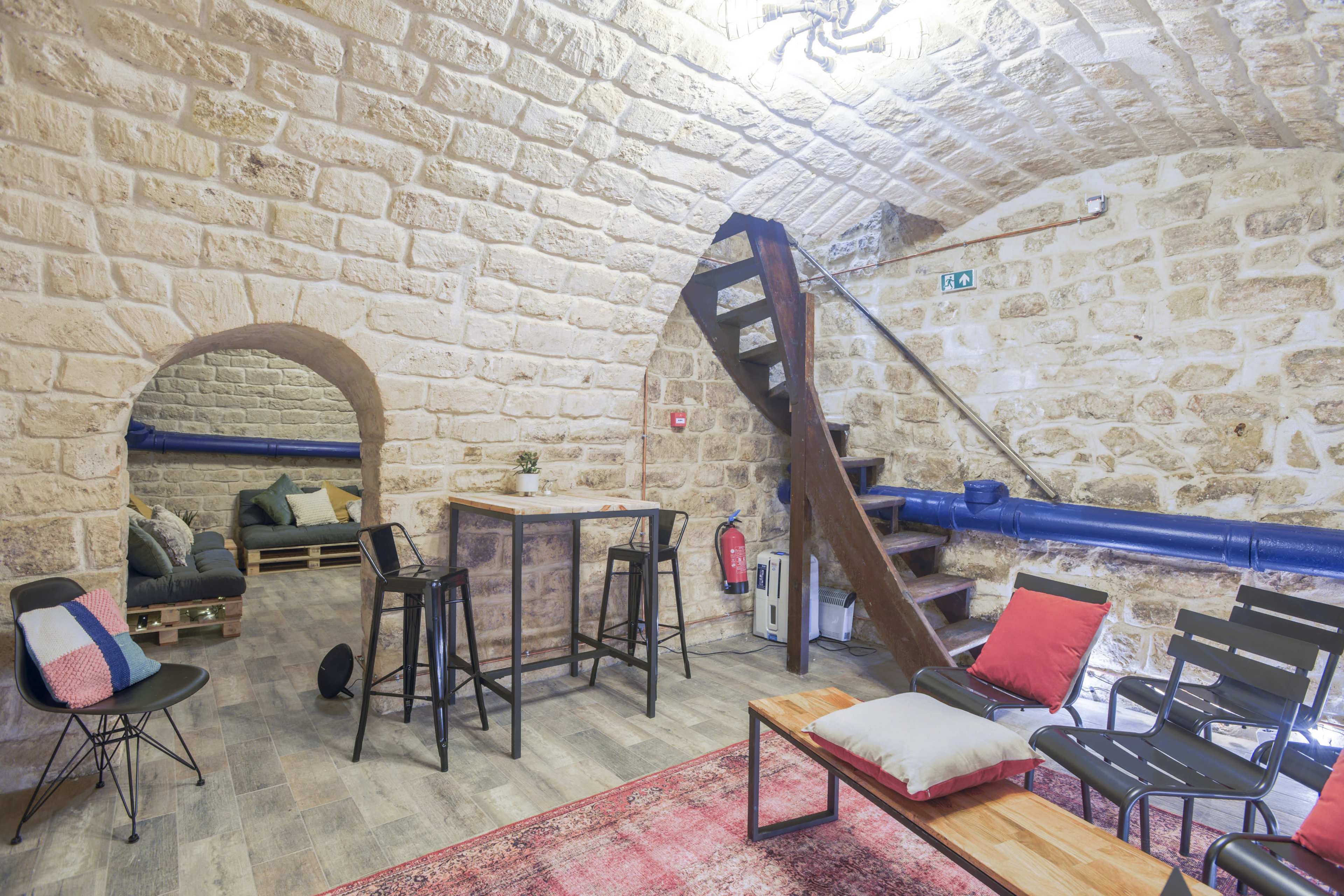 A stone-walled room featuring a small bar table with stools, a staircase, and seating areas with cushions and a rug.