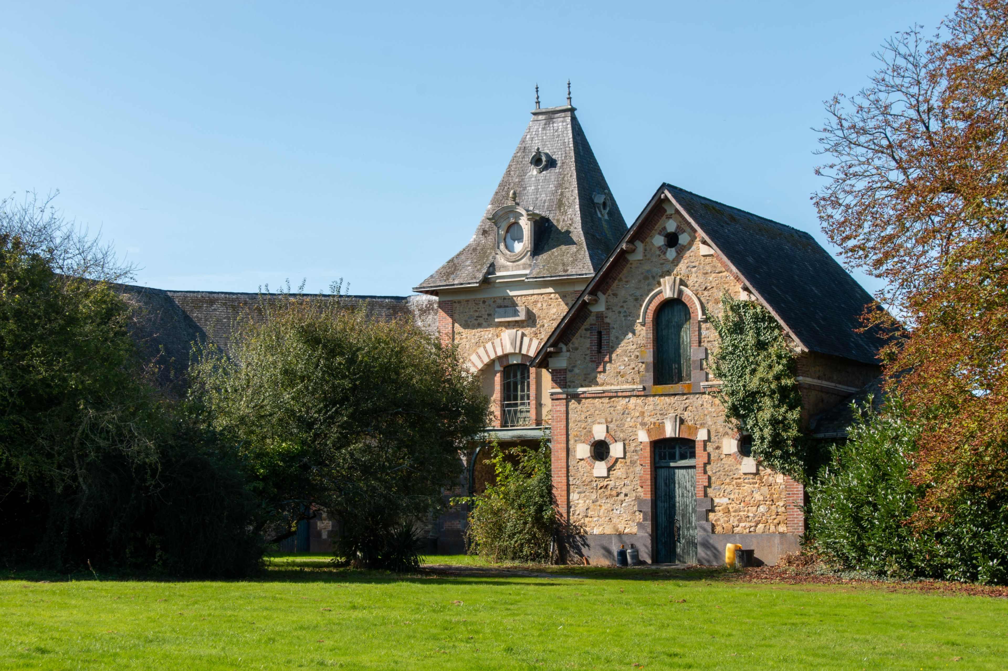 A historic stone building with a triangular roof is situated in a grassy area surrounded by trees.
