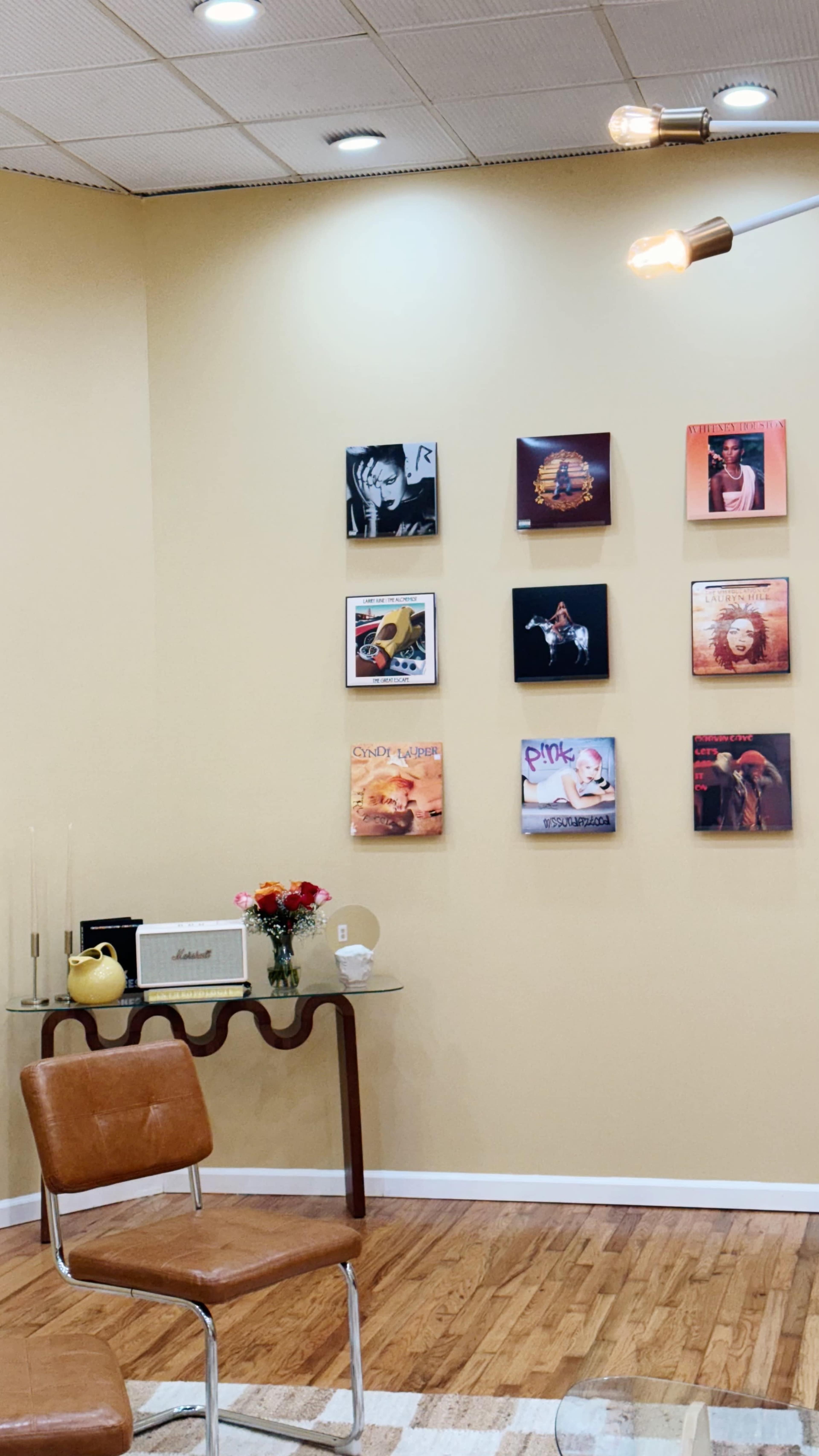 The image shows a corner of a room with a brown chair, a glass table, and a wall displaying various framed vinyl records.