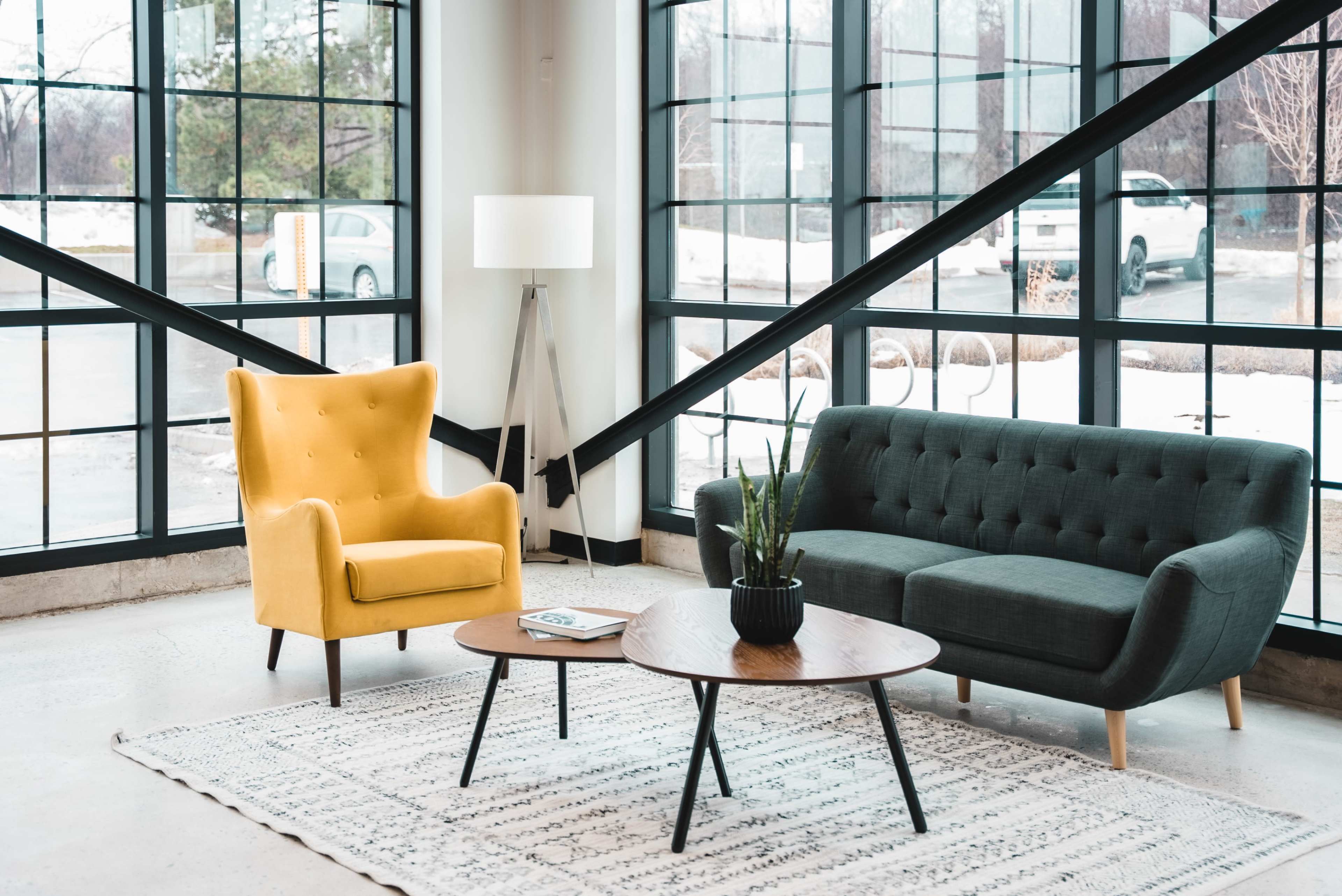A modern living area with a yellow armchair, a dark green sofa, a round coffee table, and a standing lamp, all set against large windows.