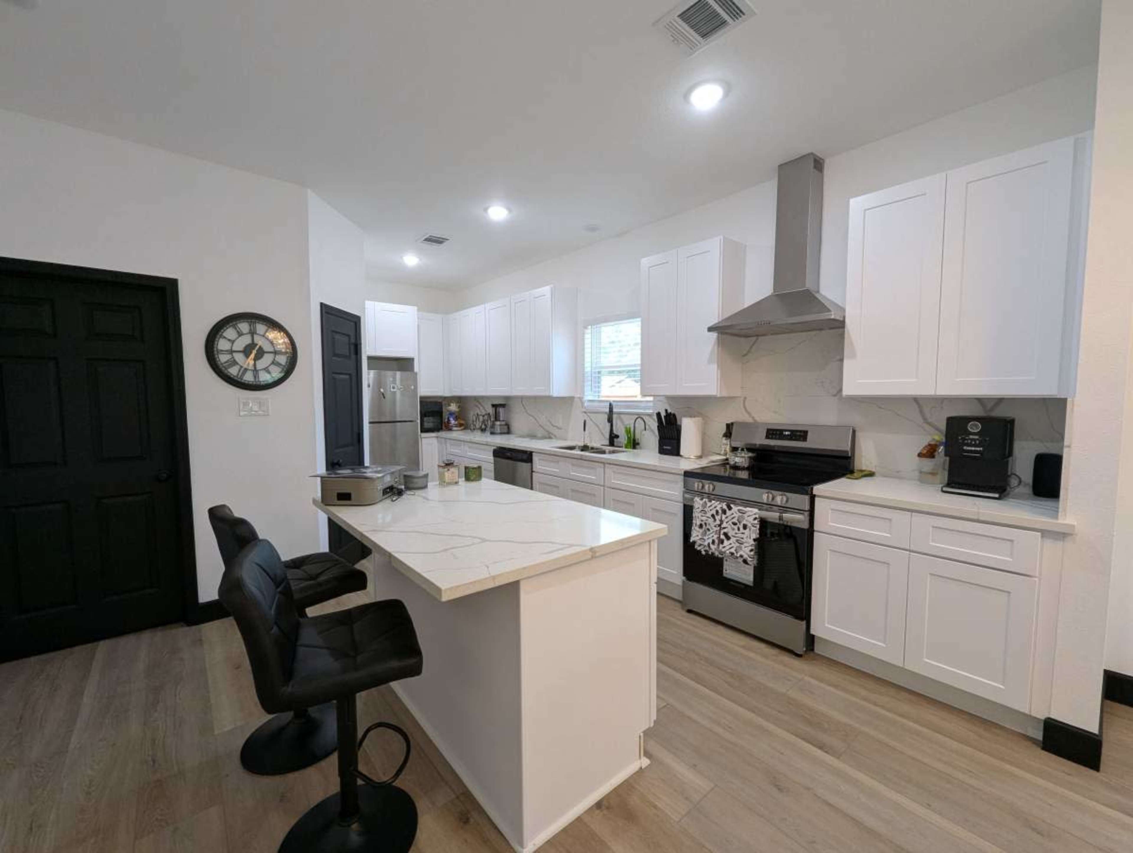 The image shows a modern kitchen featuring white cabinetry, a large island with black bar stools, stainless steel appliances, and a light-colored countertop.