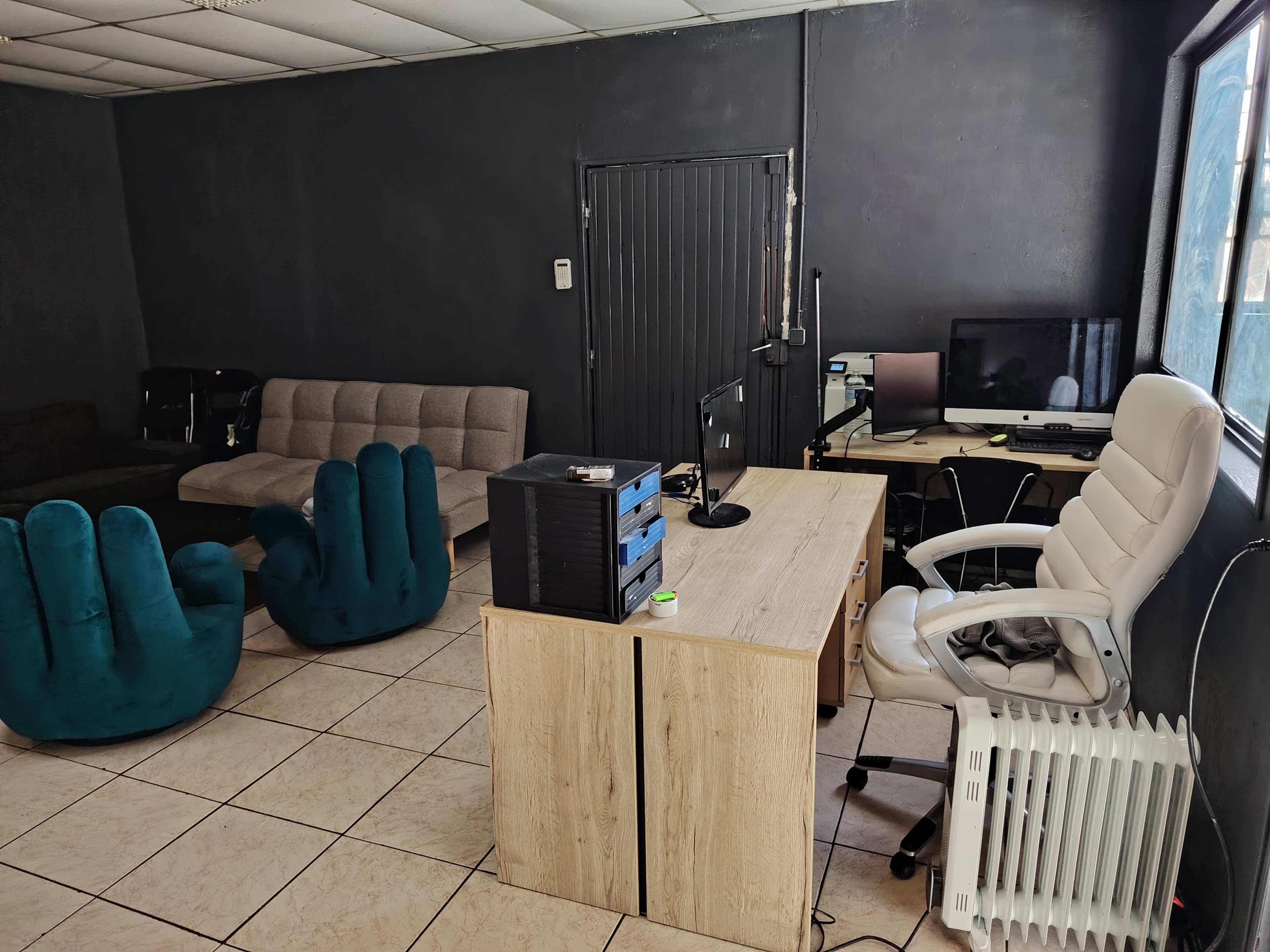 The image shows an office space featuring a wooden desk with a chair, two large blue hand-shaped chairs, a computer setup, and a portable heater against a dark wall.