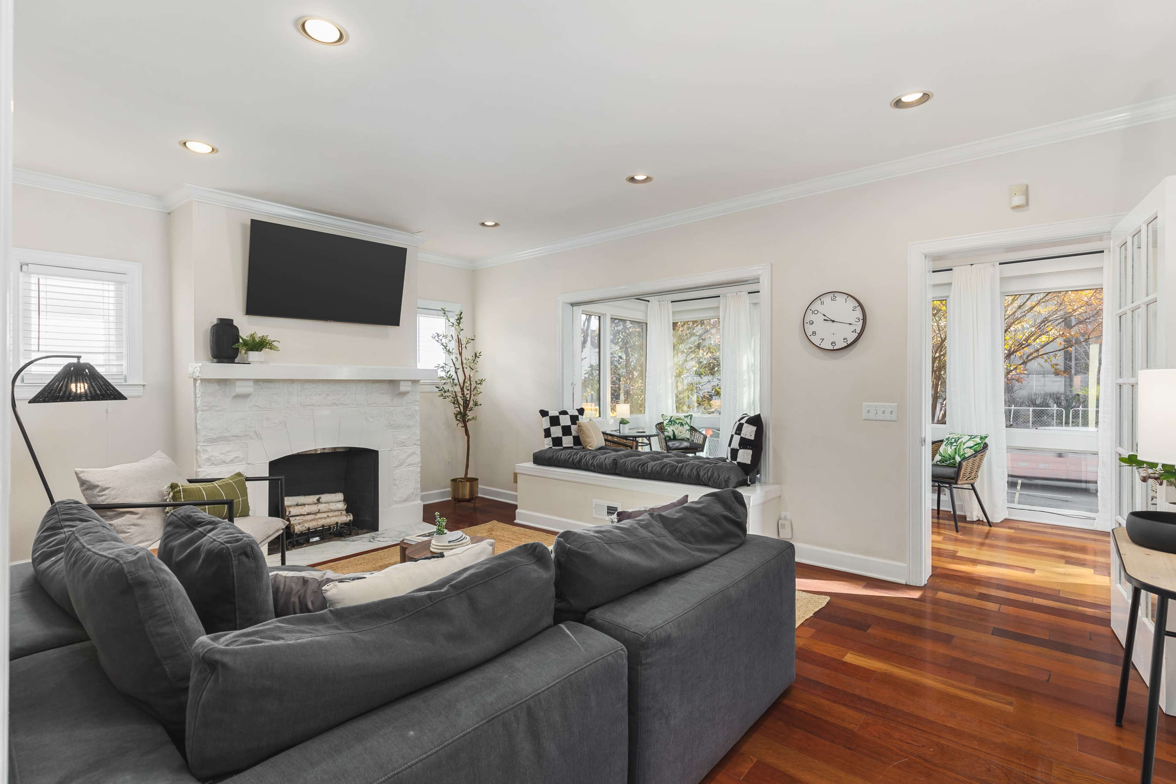 The living room features a gray sectional sofa, a white stone fireplace with a television mounted above it, and a window seat with cushions next to large windows.