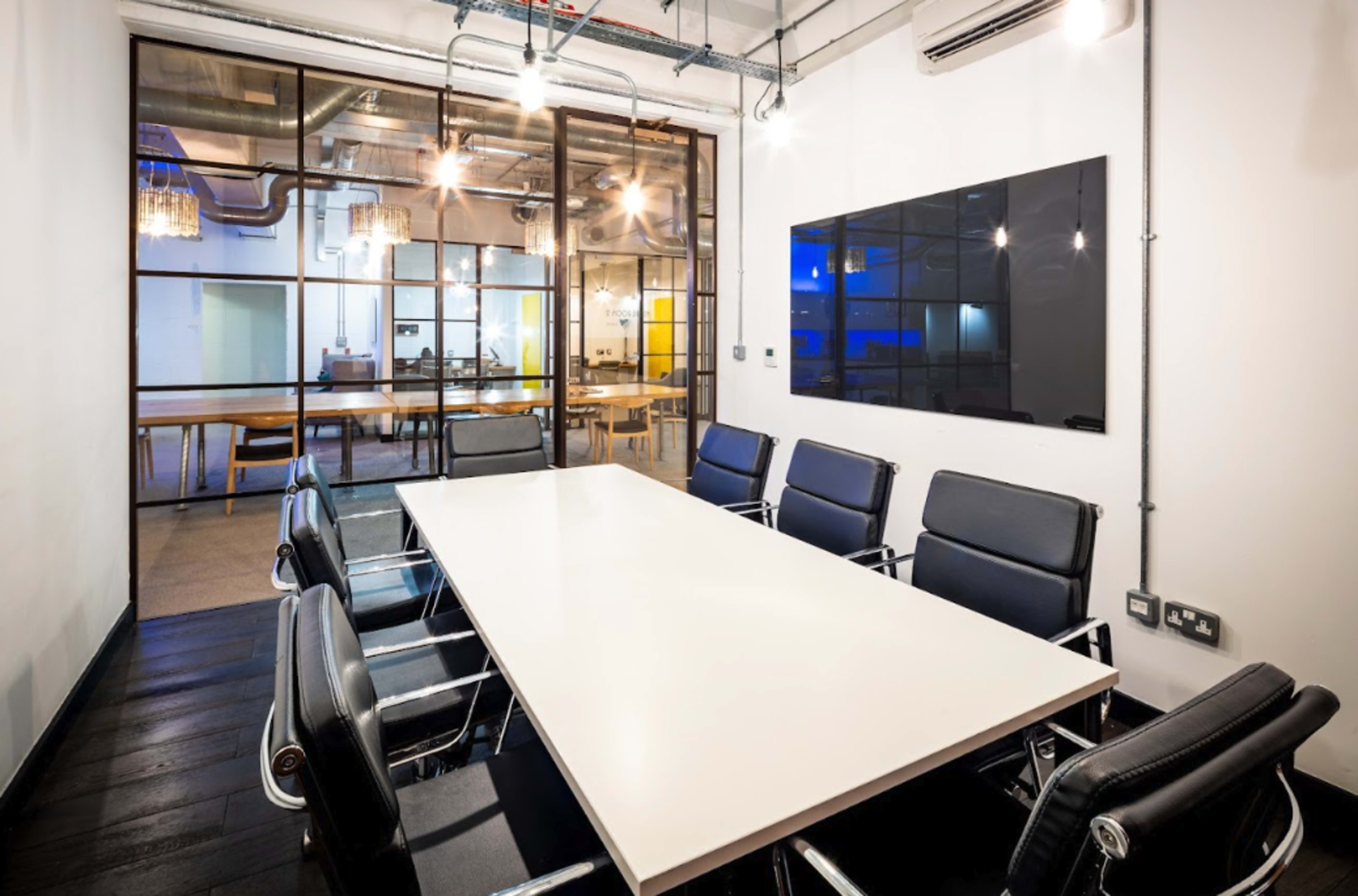 A modern conference room with a large white table surrounded by black leather chairs and a glass wall separating it from another workspace.
