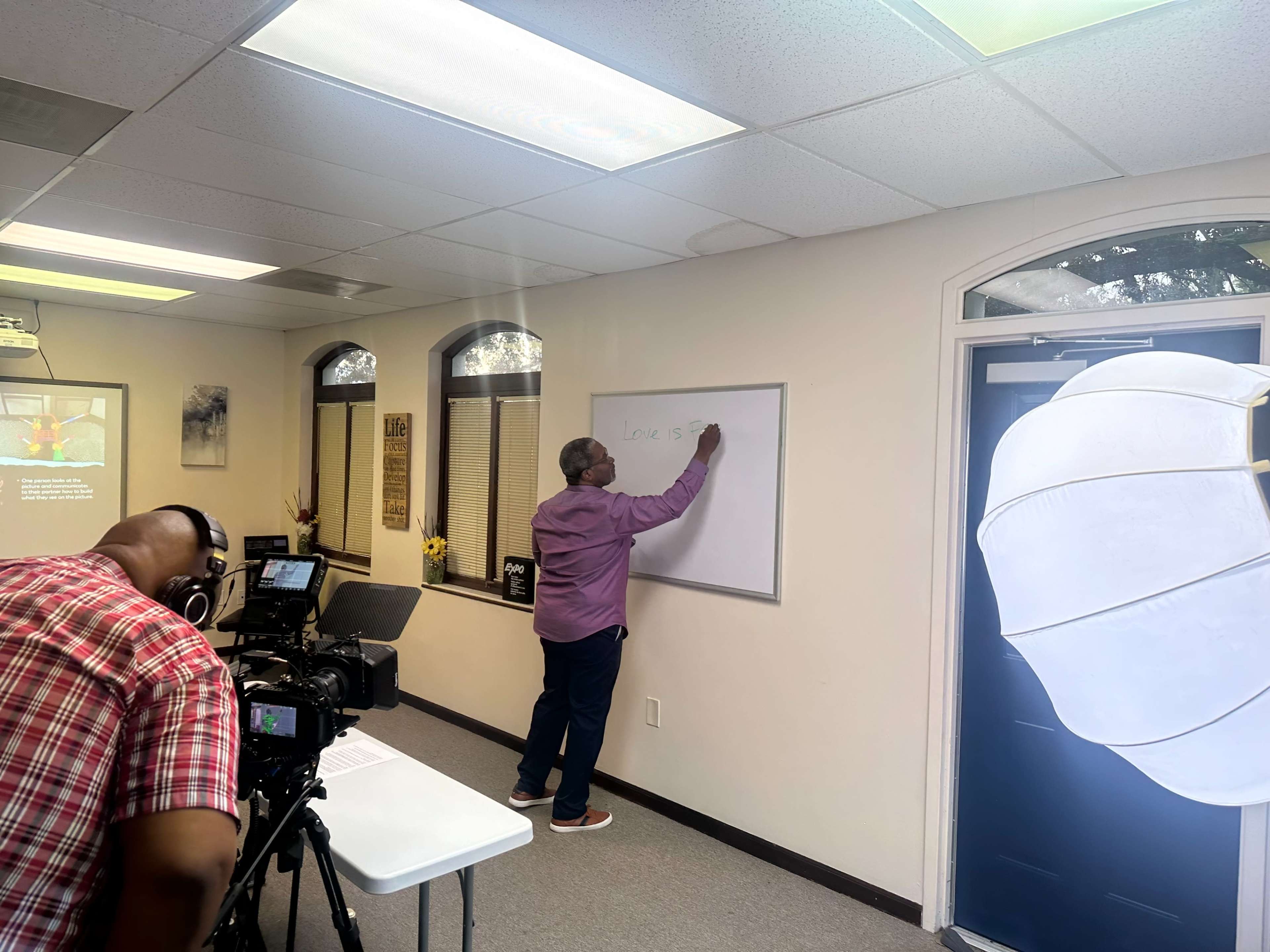 A person is writing on a whiteboard while another individual operates a camera in a brightly lit room with a table and a backdrop.