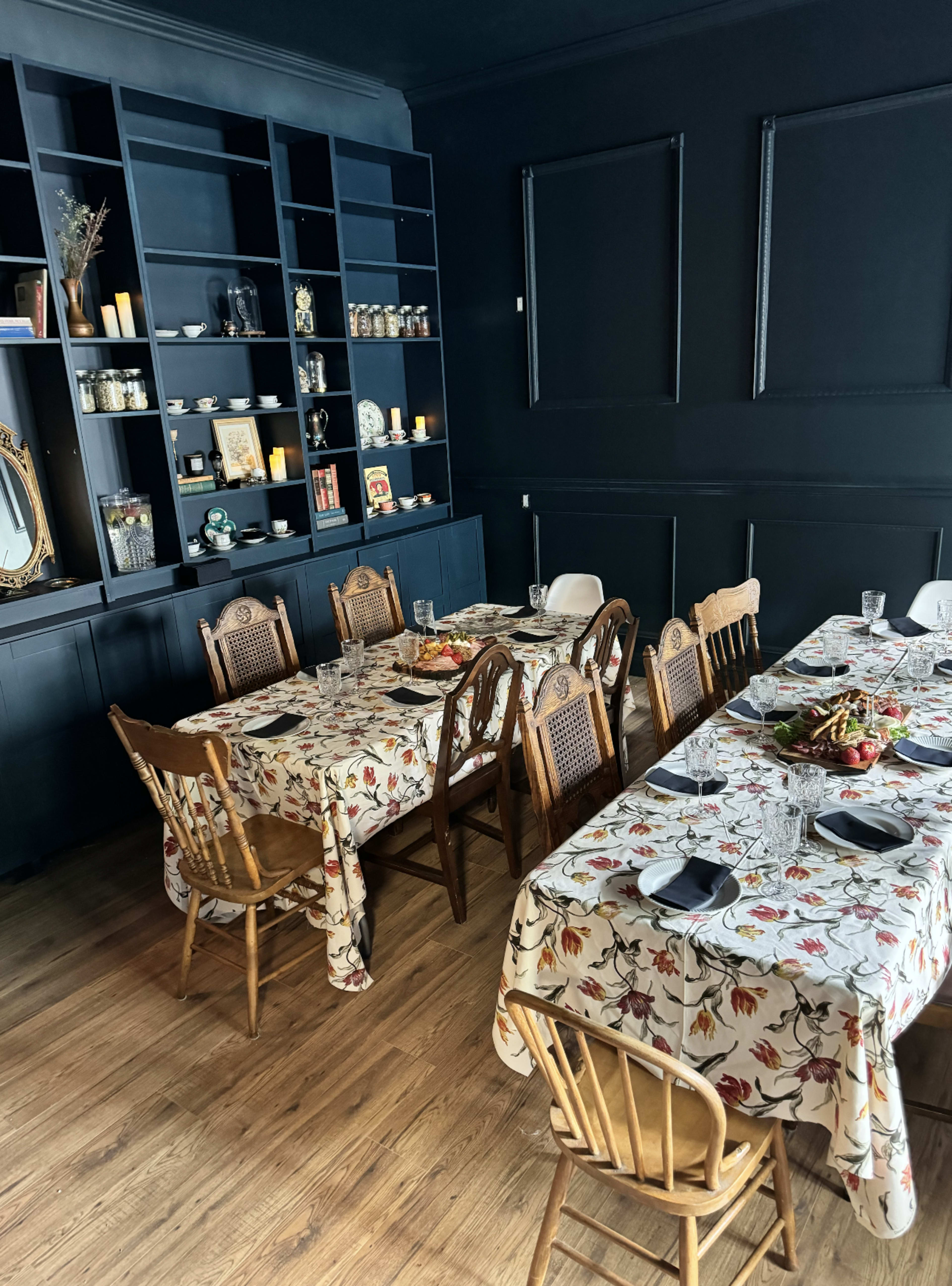 A dining area features two long tables with floral tablecloths and wooden chairs, set against a black wall lined with shelves displaying various decor items.
