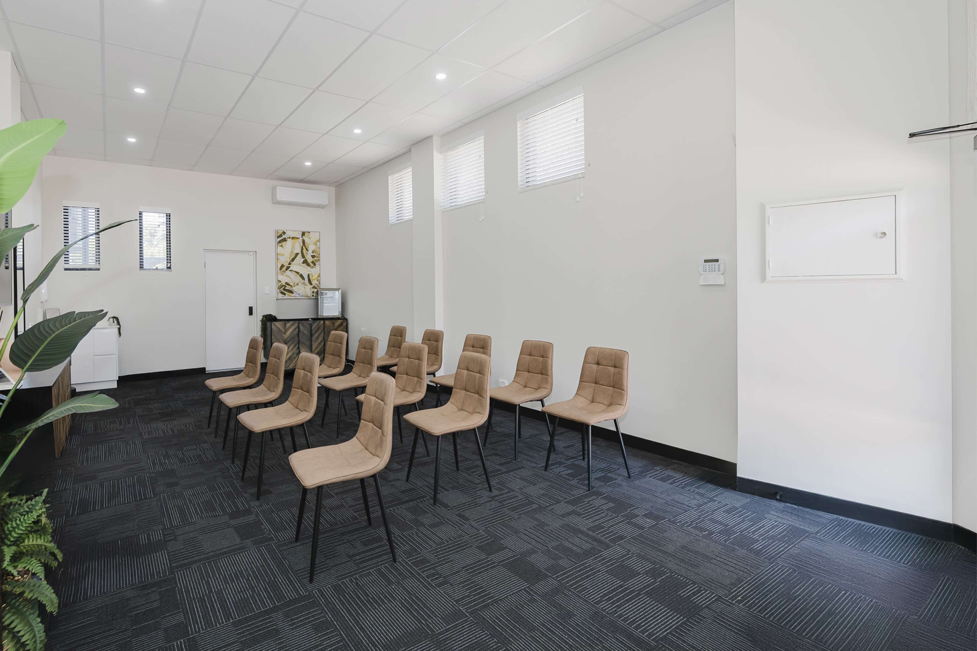 The image shows a room with a row of beige chairs arranged neatly on a black carpet, facing a white wall, with windows providing natural light.