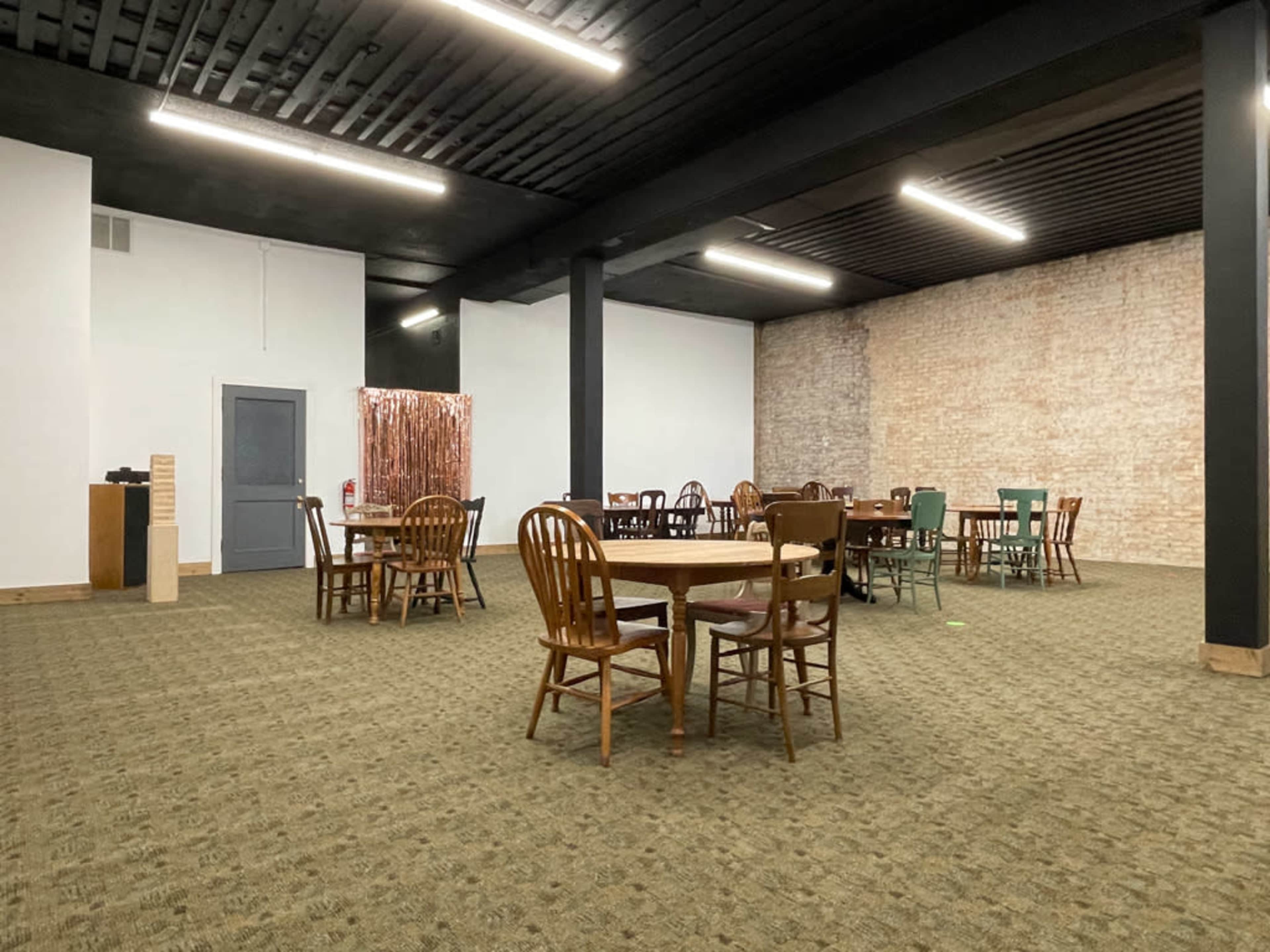 A spacious, empty room with several wooden tables and chairs arranged throughout, featuring a textured brick wall and dark ceiling.