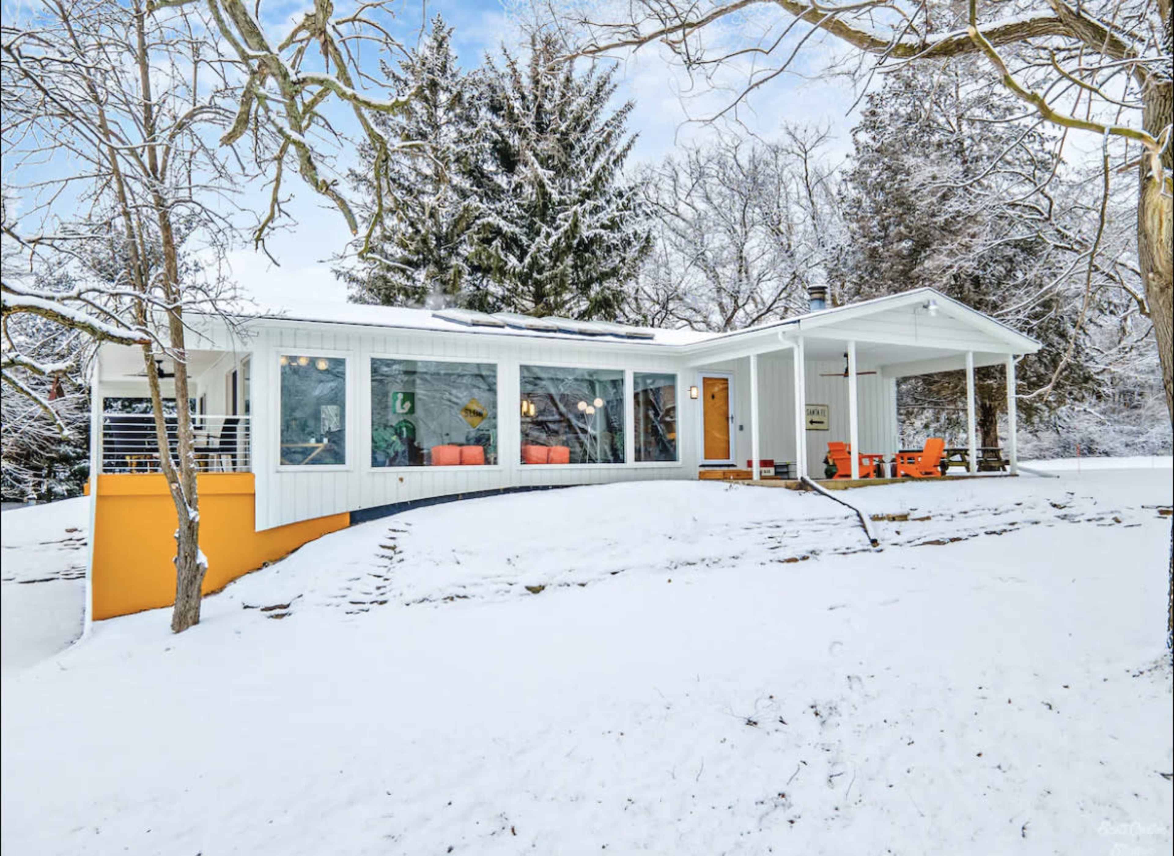 A modern house with large windows and a covered porch is surrounded by snow-covered ground and trees.