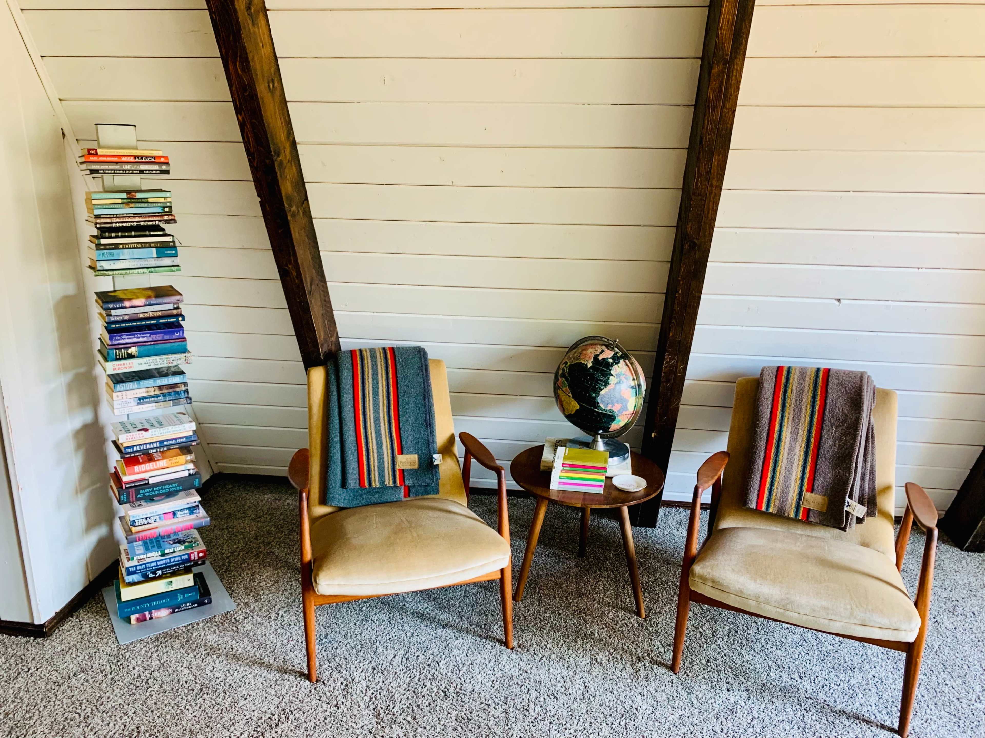 The image shows a cozy reading nook with two chairs, a small table, a globe, and a tall stack of books against a white, slanted wall.