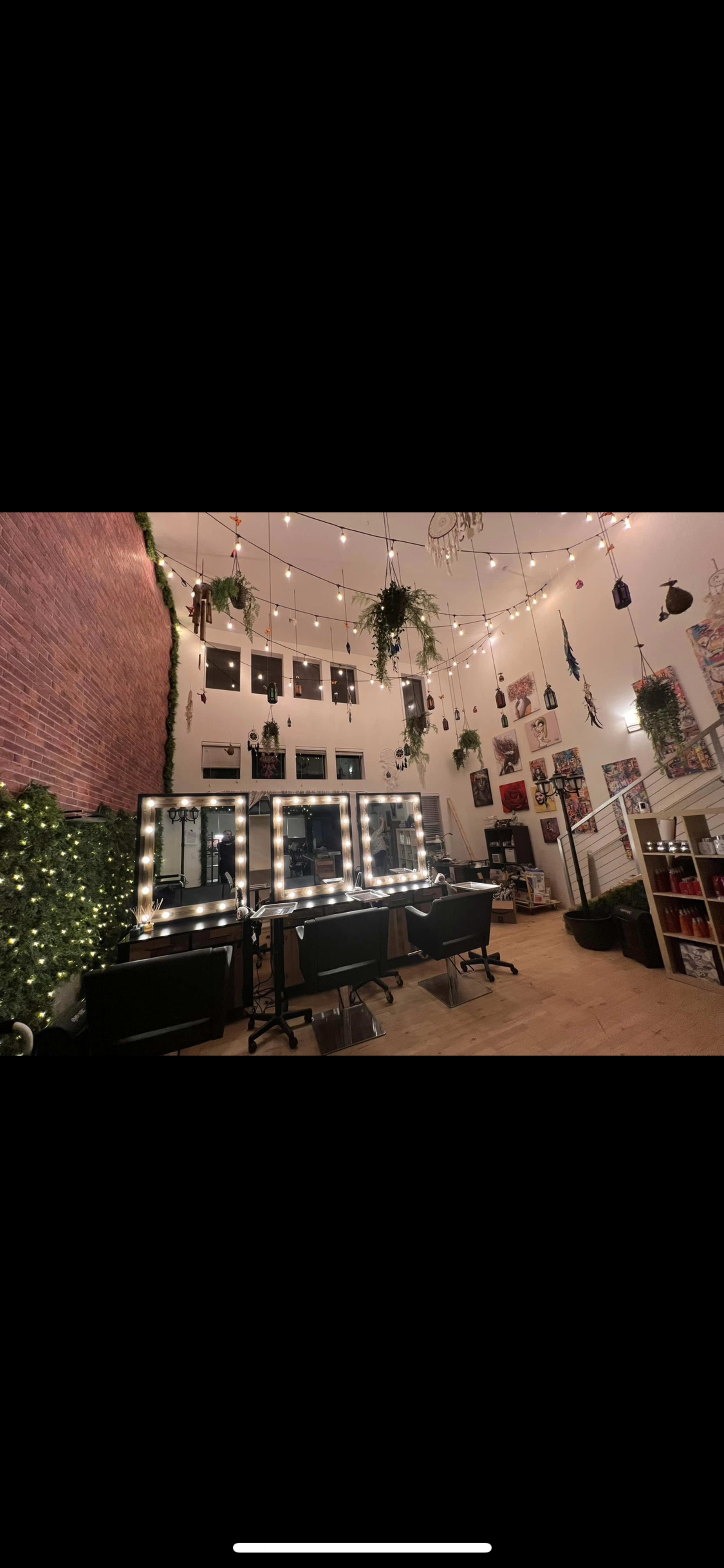 The image shows a spacious room with a brick wall, multiple mirrors surrounded by lights, hanging plants, and several chairs arranged for use.
