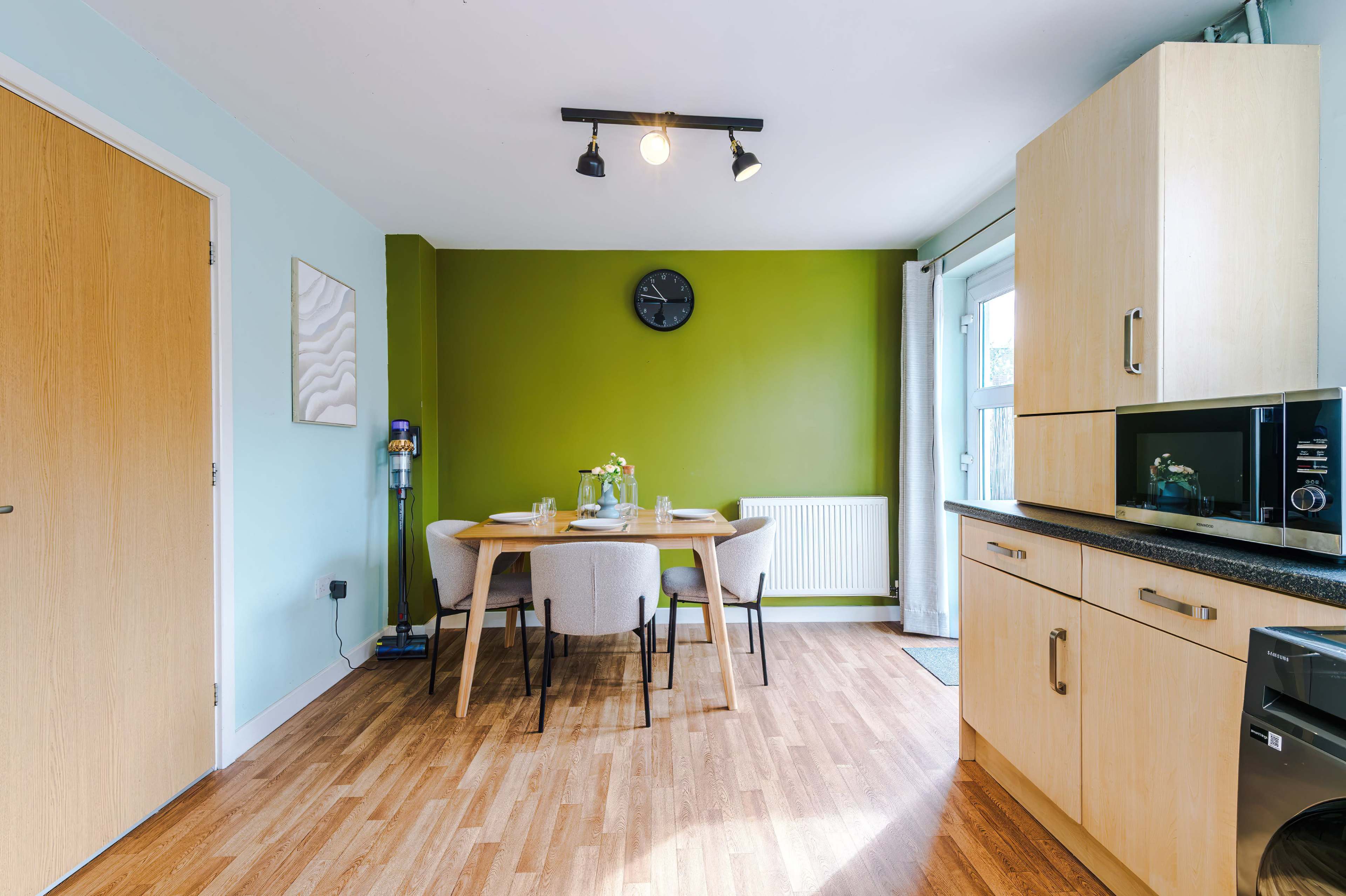 A modern kitchen features a dining table set for four, with a green accent wall and wooden flooring.