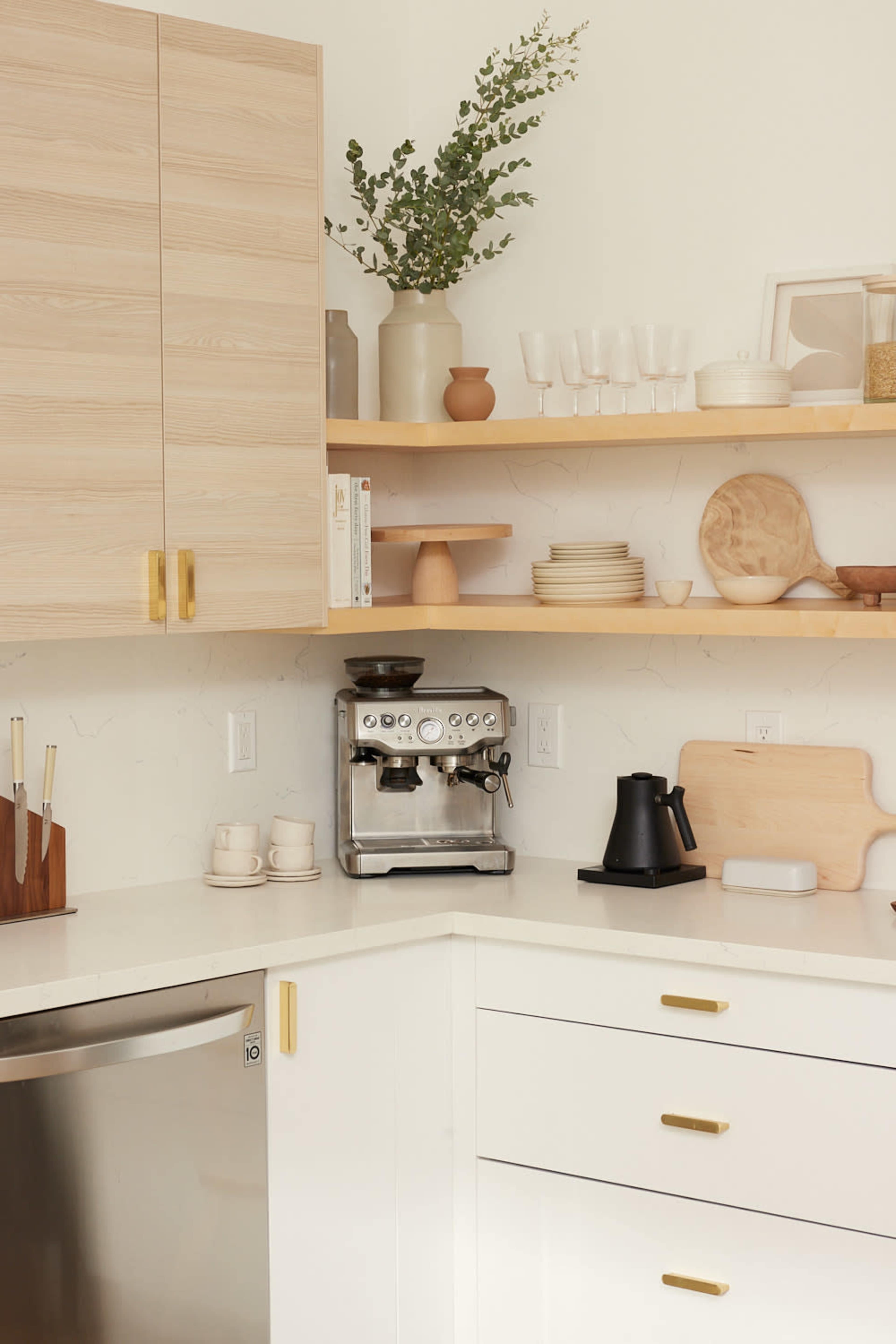 The image shows a modern kitchen with light wood cabinets, open shelves displaying dishware, and a coffee machine on the countertop.
