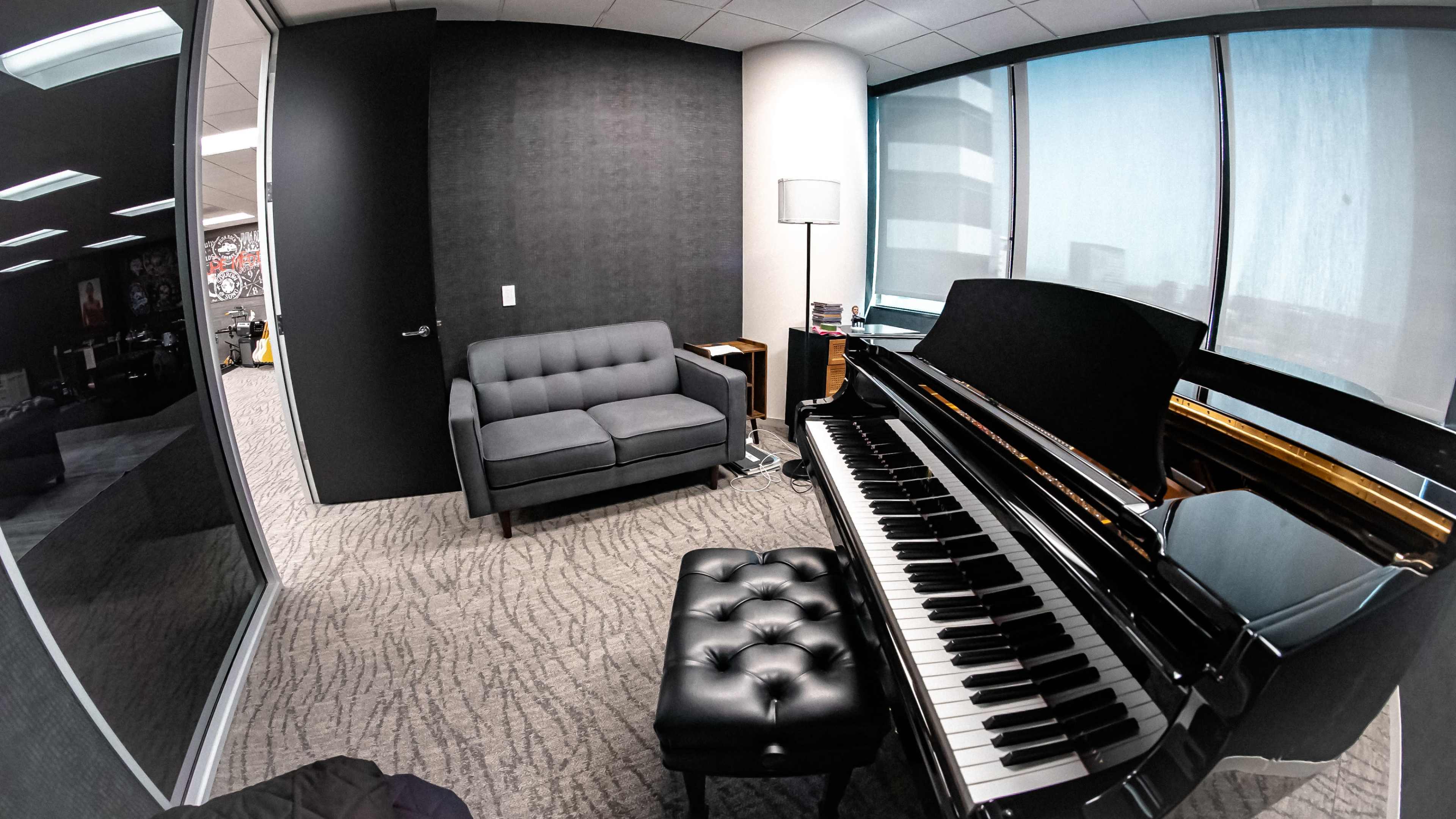 A corner of an office space featuring a black piano, a gray couch, a lamp, and large windows.