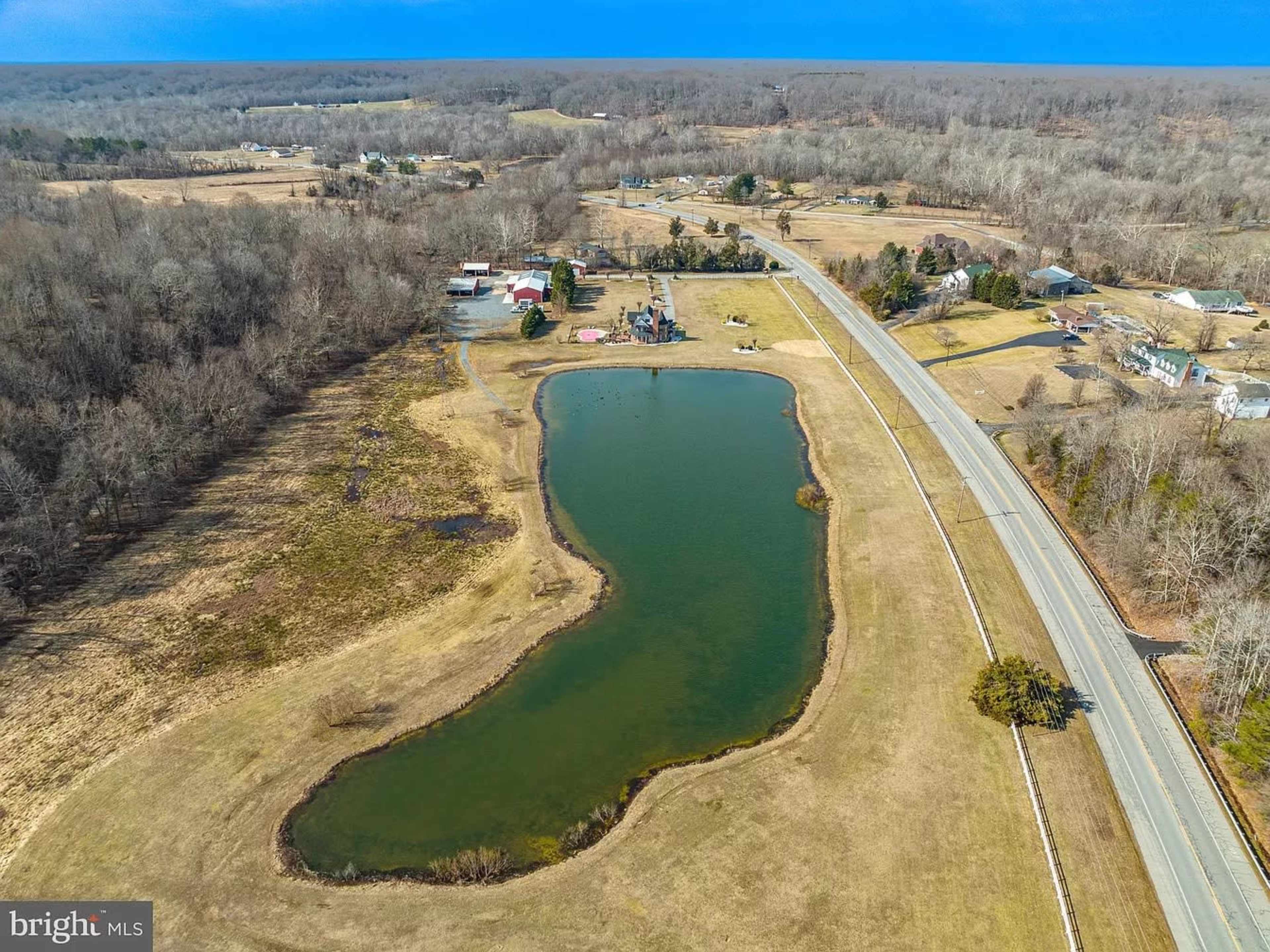 An aerial view of a curved pond next to a road, surrounded by open fields and scattered buildings.