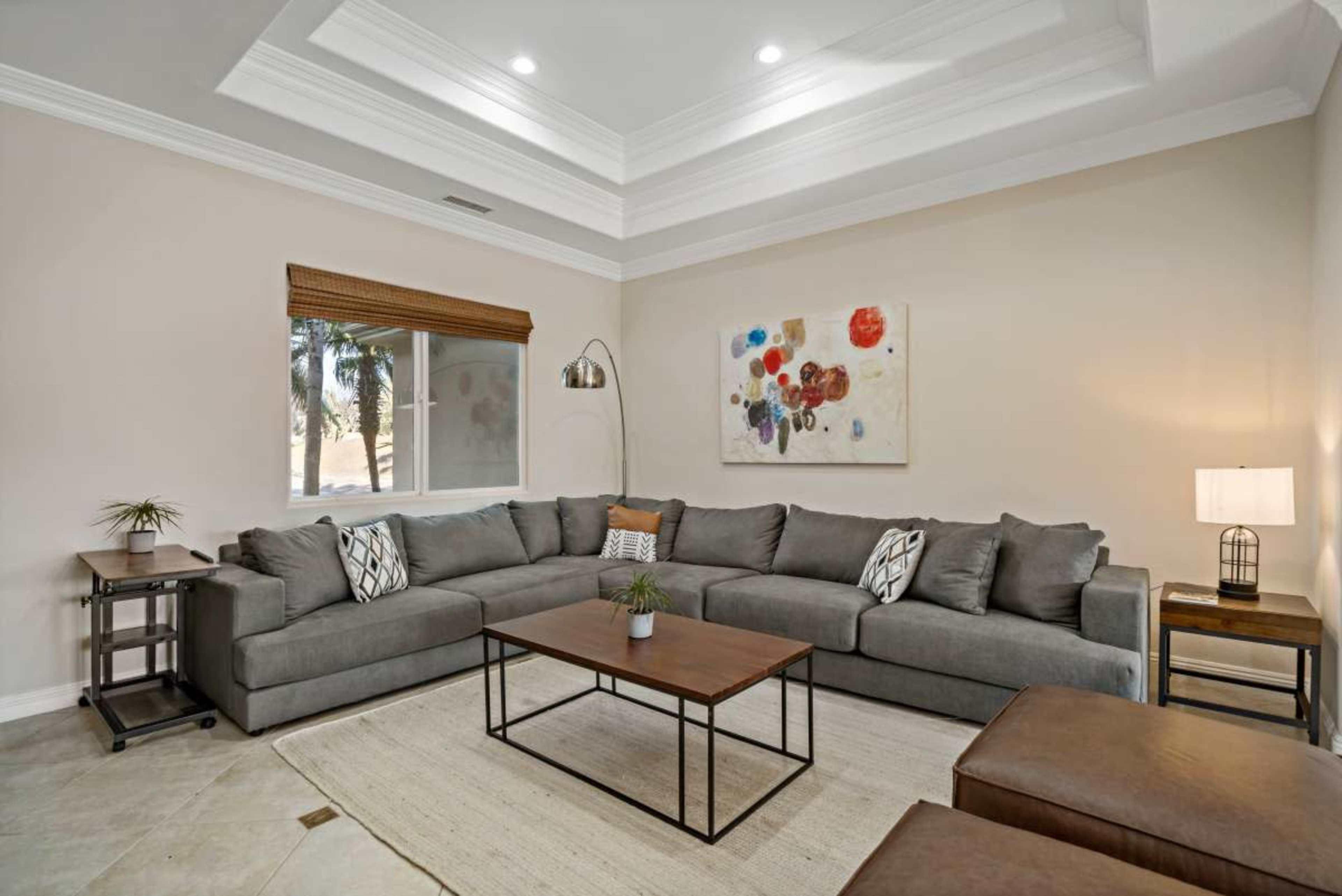 The image shows a modern living room featuring a large gray sectional sofa, a coffee table, and a lamp, with a window displaying a view of palm trees in the background.