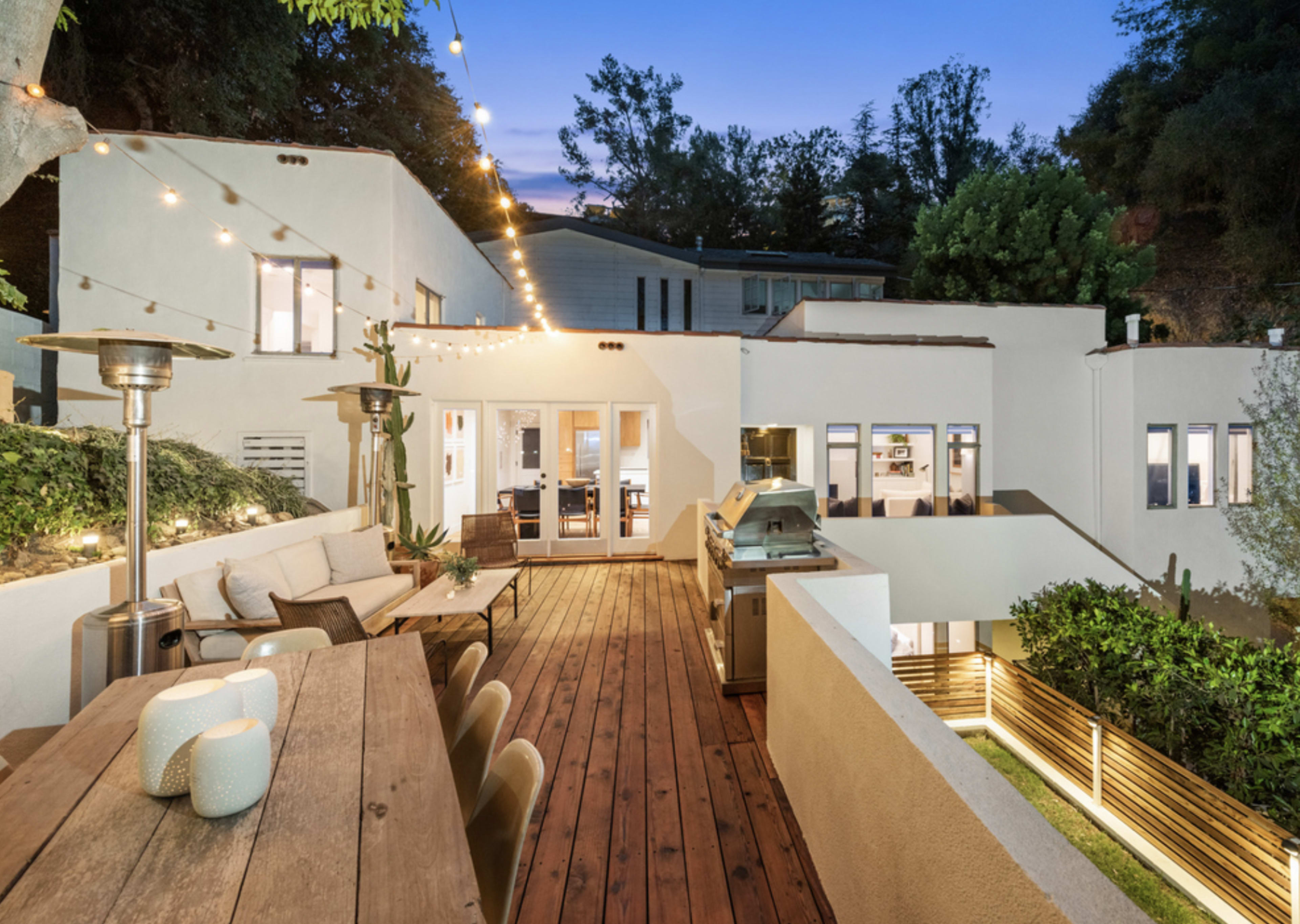 A modern home features a spacious deck with a dining area, outdoor heaters, and string lights, surrounded by greenery and a barbecue area.