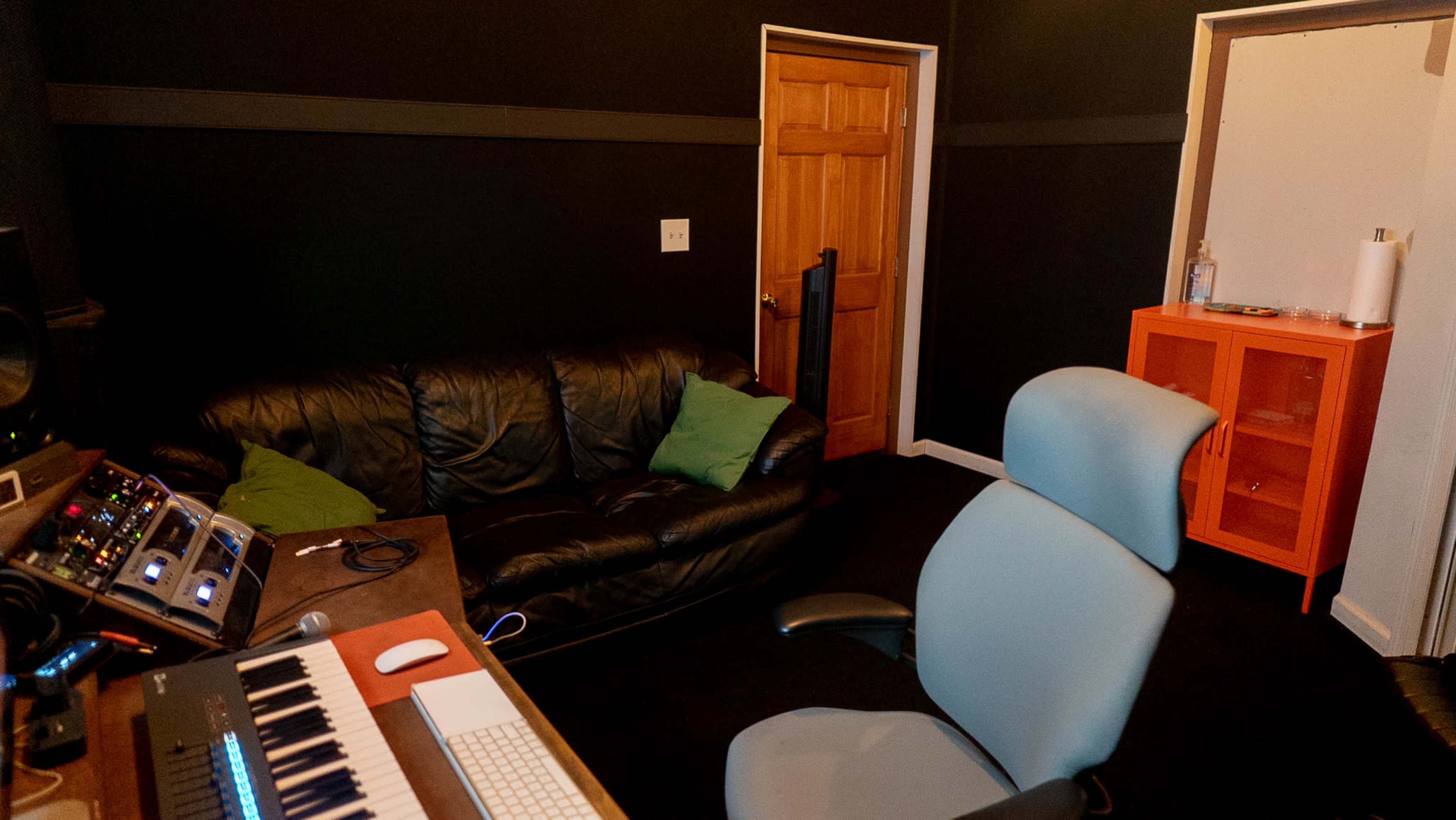 The image shows a small recording studio with a black leather couch, a desk featuring audio equipment, and an orange cabinet against a dark wall.