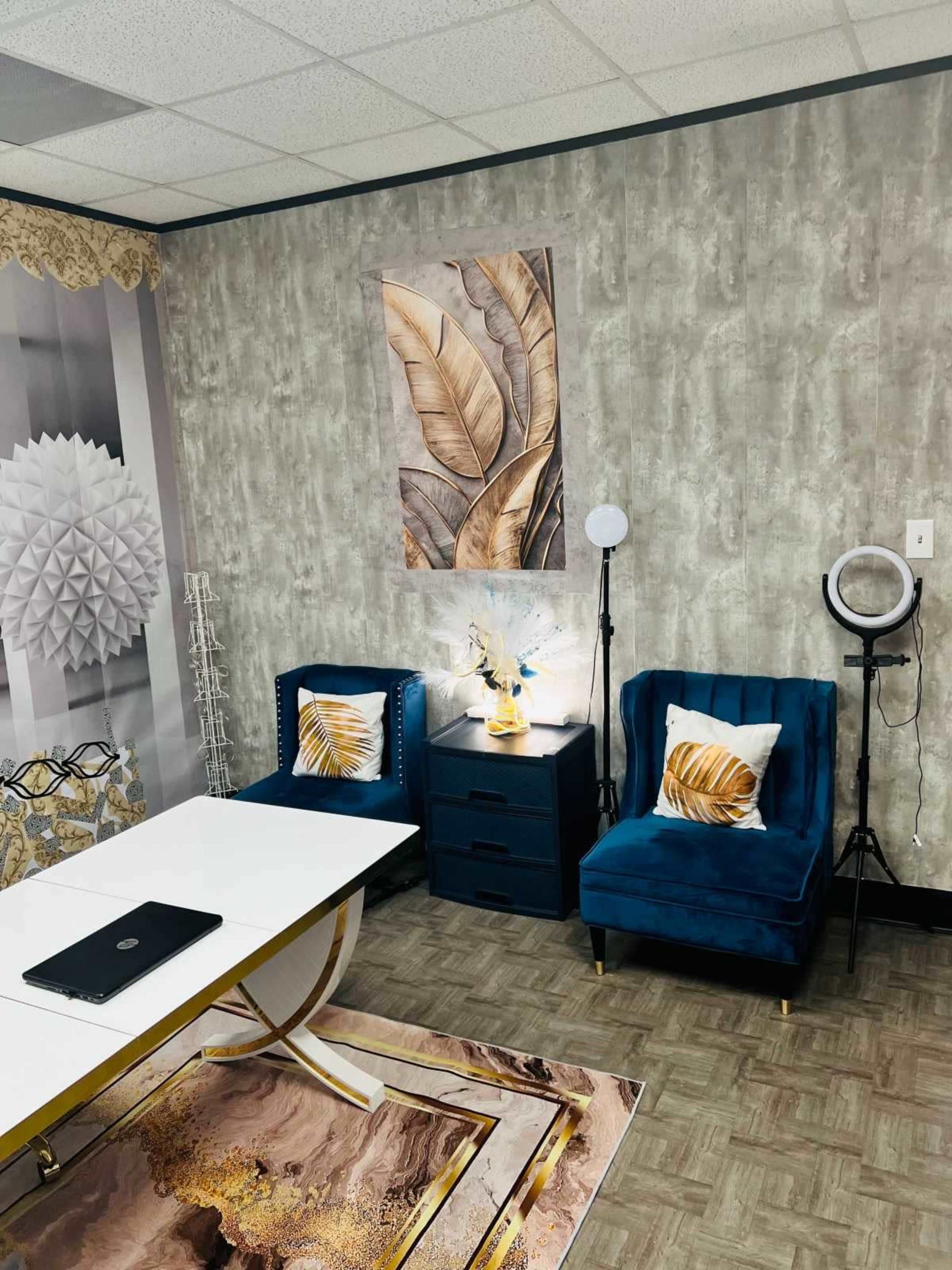 The image shows a stylish office interior featuring two blue velvet chairs, a dark bedside table, a large wall art piece with leaf designs, and a lighted mirror on a stand.