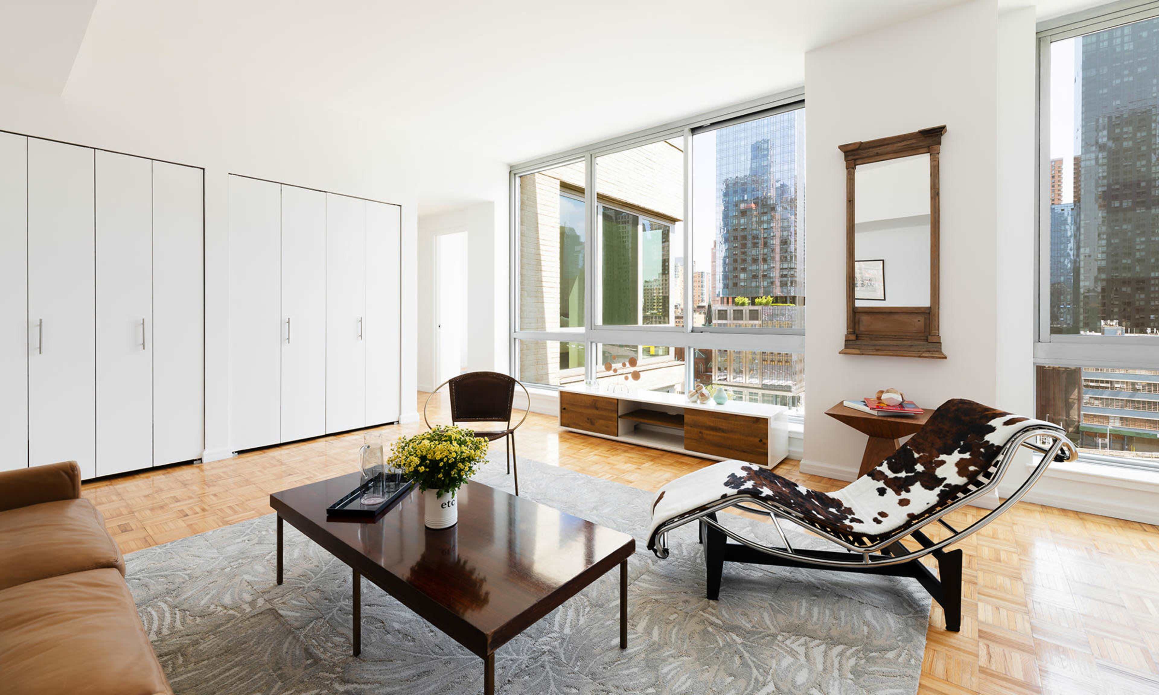 The image shows a modern living room featuring a brown sofa, a sleek coffee table, a patterned area rug, and large windows with views of a cityscape.