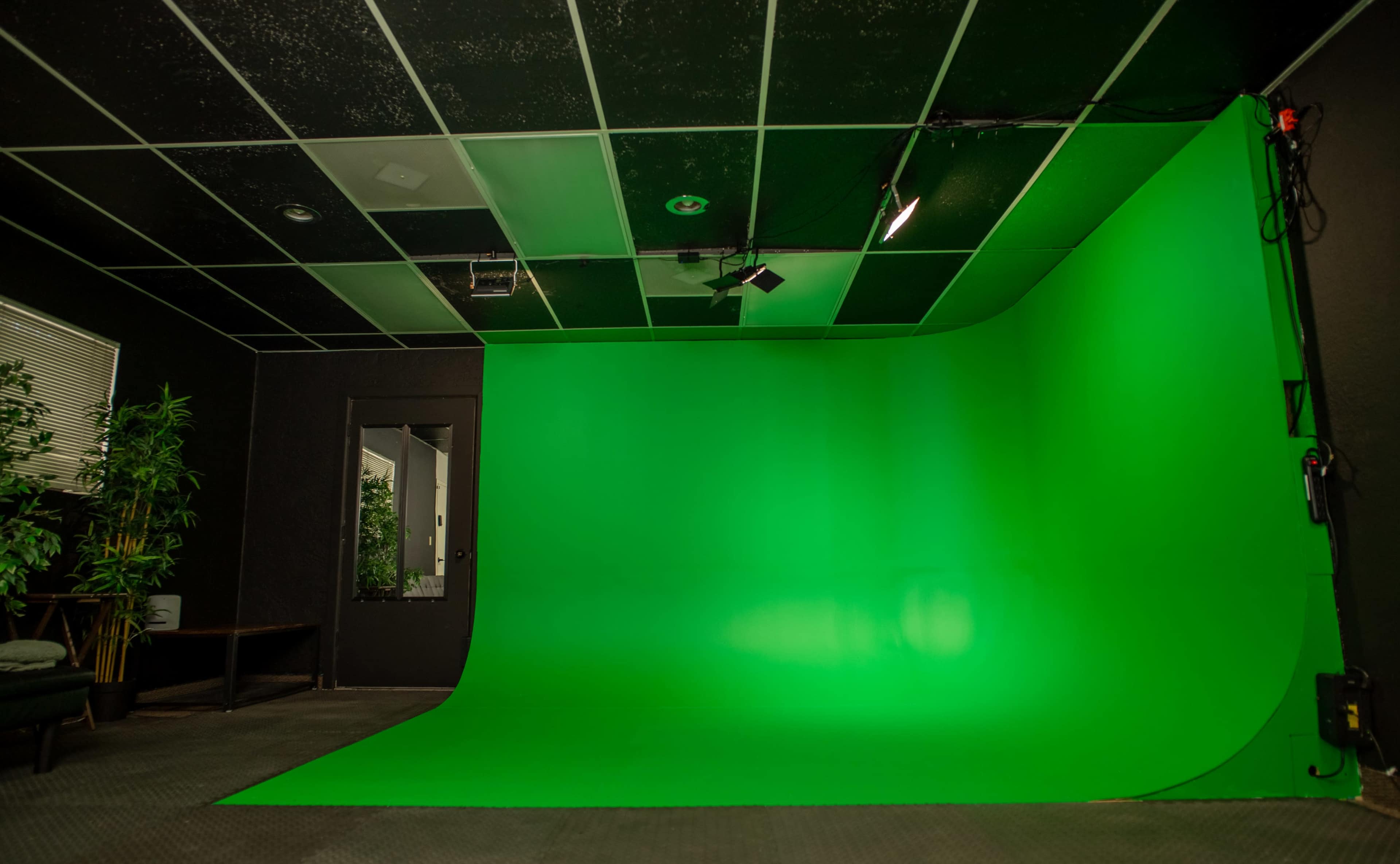 The image shows a green screen setup in a room with black walls and a ceiling, featuring a smooth green backdrop and minimal furnishings.