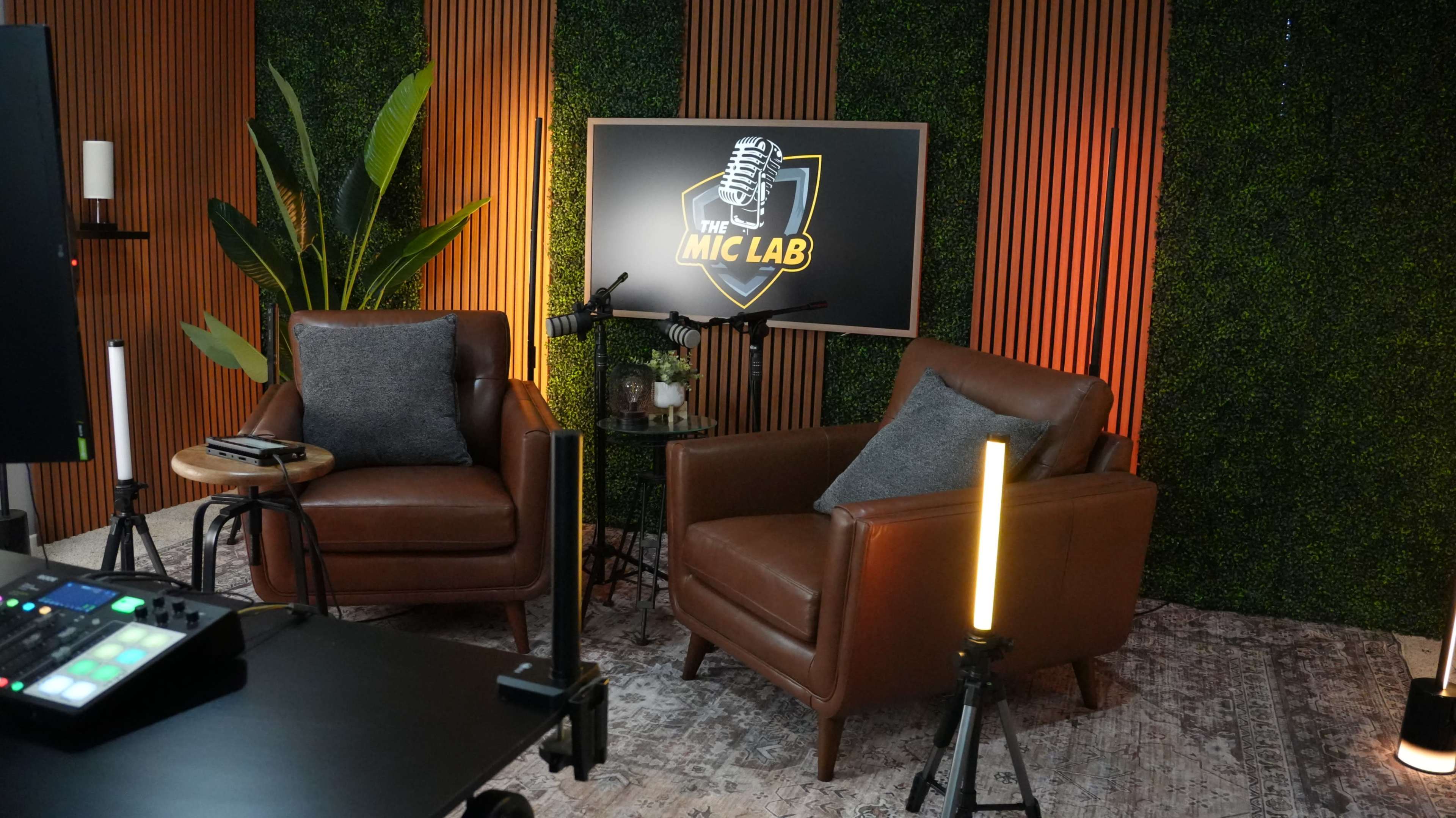 The image shows a cozy studio setup featuring two brown leather chairs, a microphone, a decorative plant, and a large screen displaying "The Mic Lab" on a green wall backdrop.