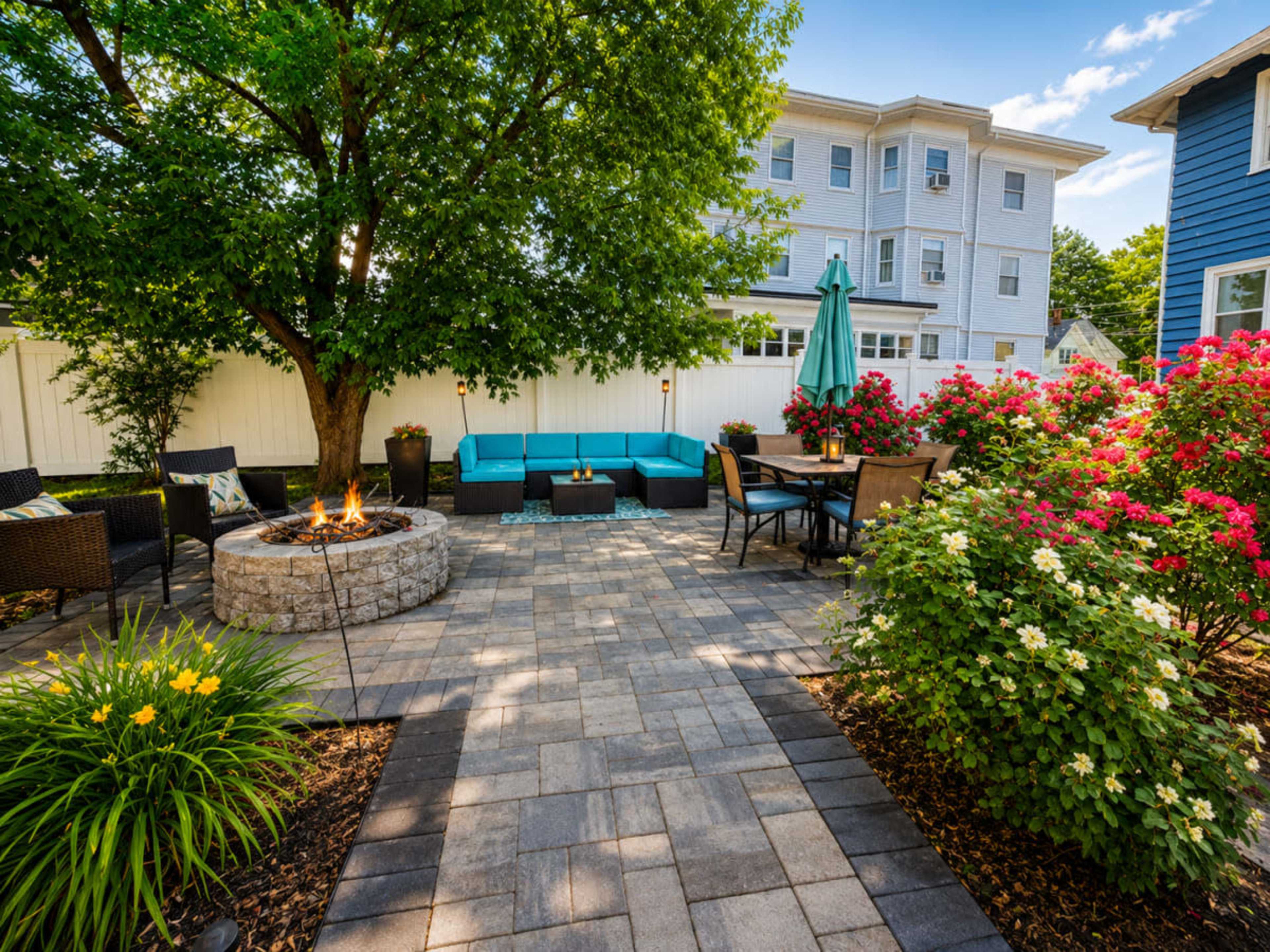 A backyard patio features a stone fire pit surrounded by seating, with a blue couch, dining area, and blooming flowers.