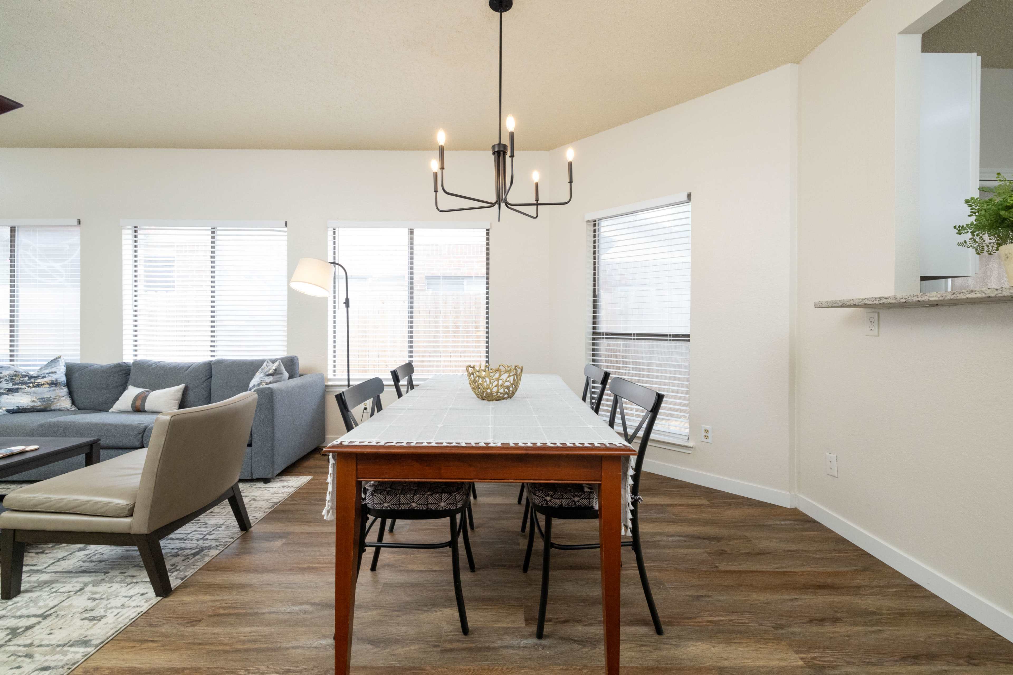 The image shows a modern dining area with a long wooden table, black chairs, and a chandelier, adjacent to a living space with a gray couch and decorative lamp.