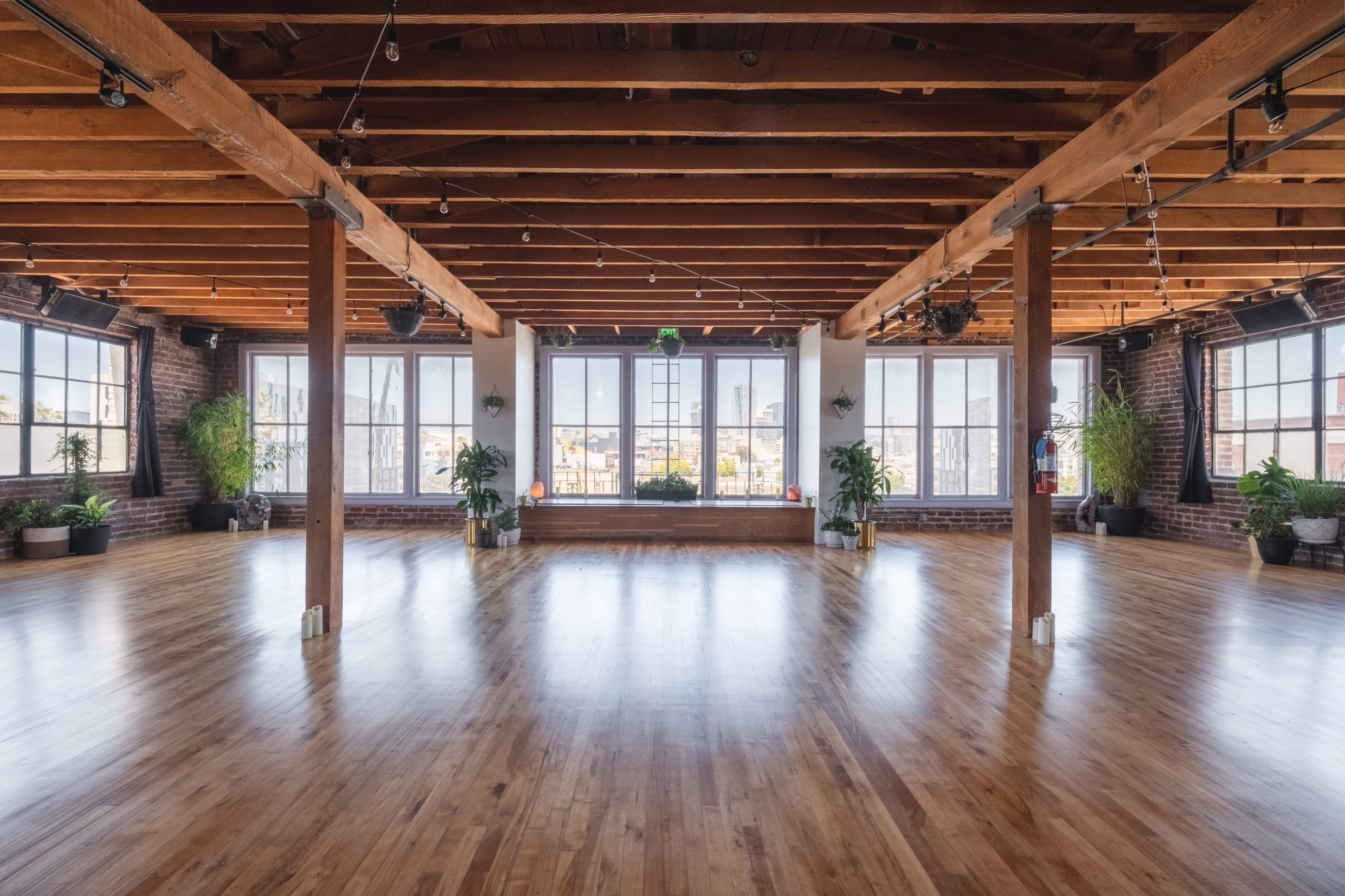A spacious, empty room with wooden floors and exposed beams features large windows and green plants along the walls.