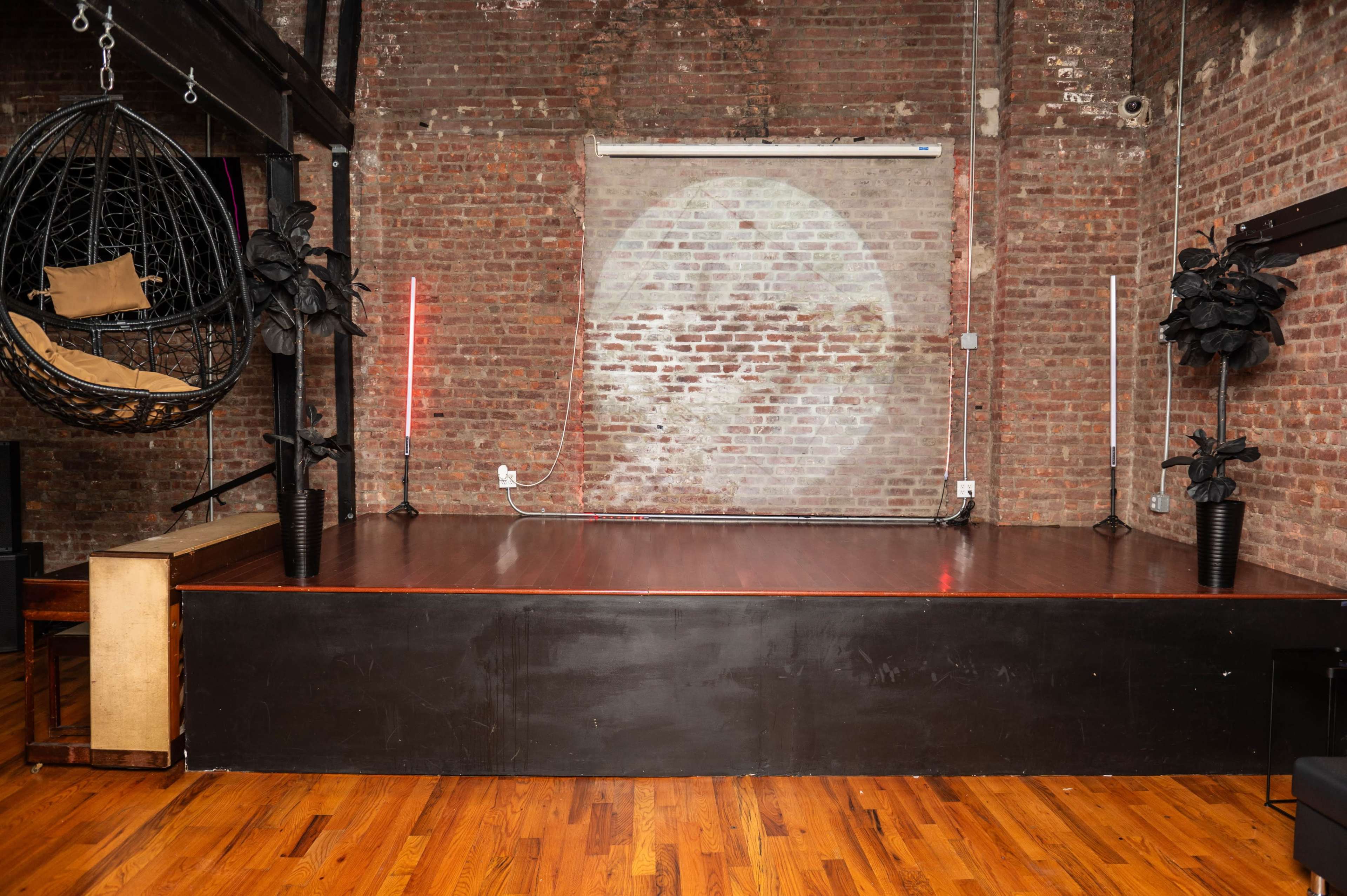 The image shows a dimly lit room with exposed brick walls, a wooden floor, a raised stage area, and a hanging chair alongside potted plants.