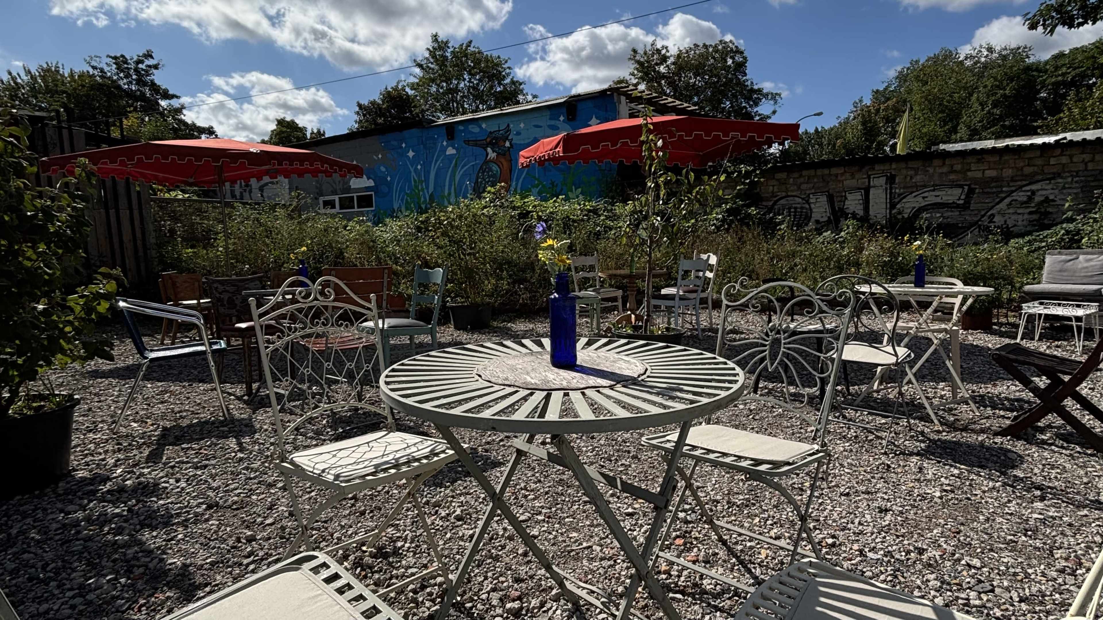 The image shows a small outdoor courtyard with white metal tables and chairs, surrounded by greenery and featuring a colorful mural on a nearby wall.