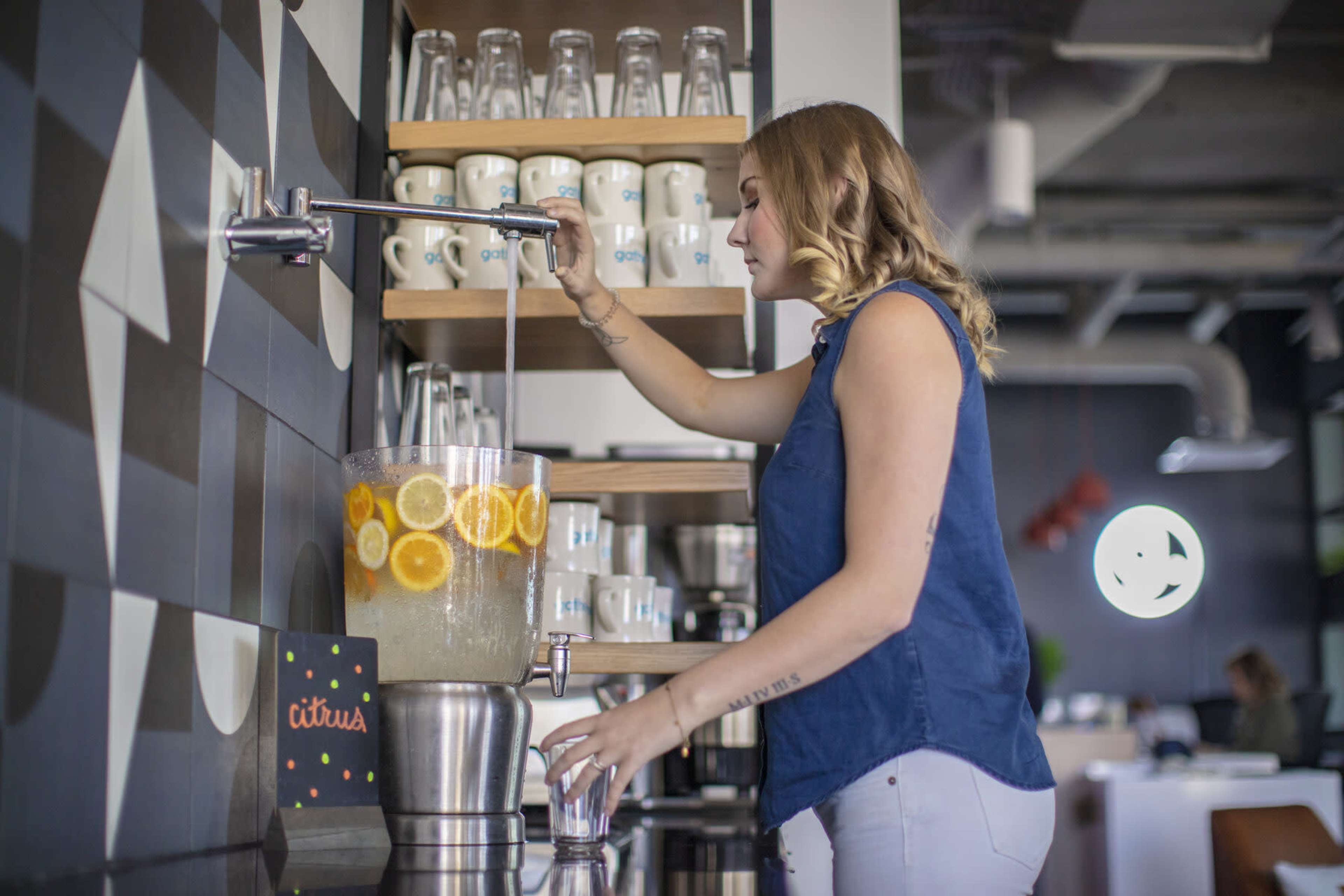 A woman is dispensing a citrus beverage from a large glass dispenser in a cafe.