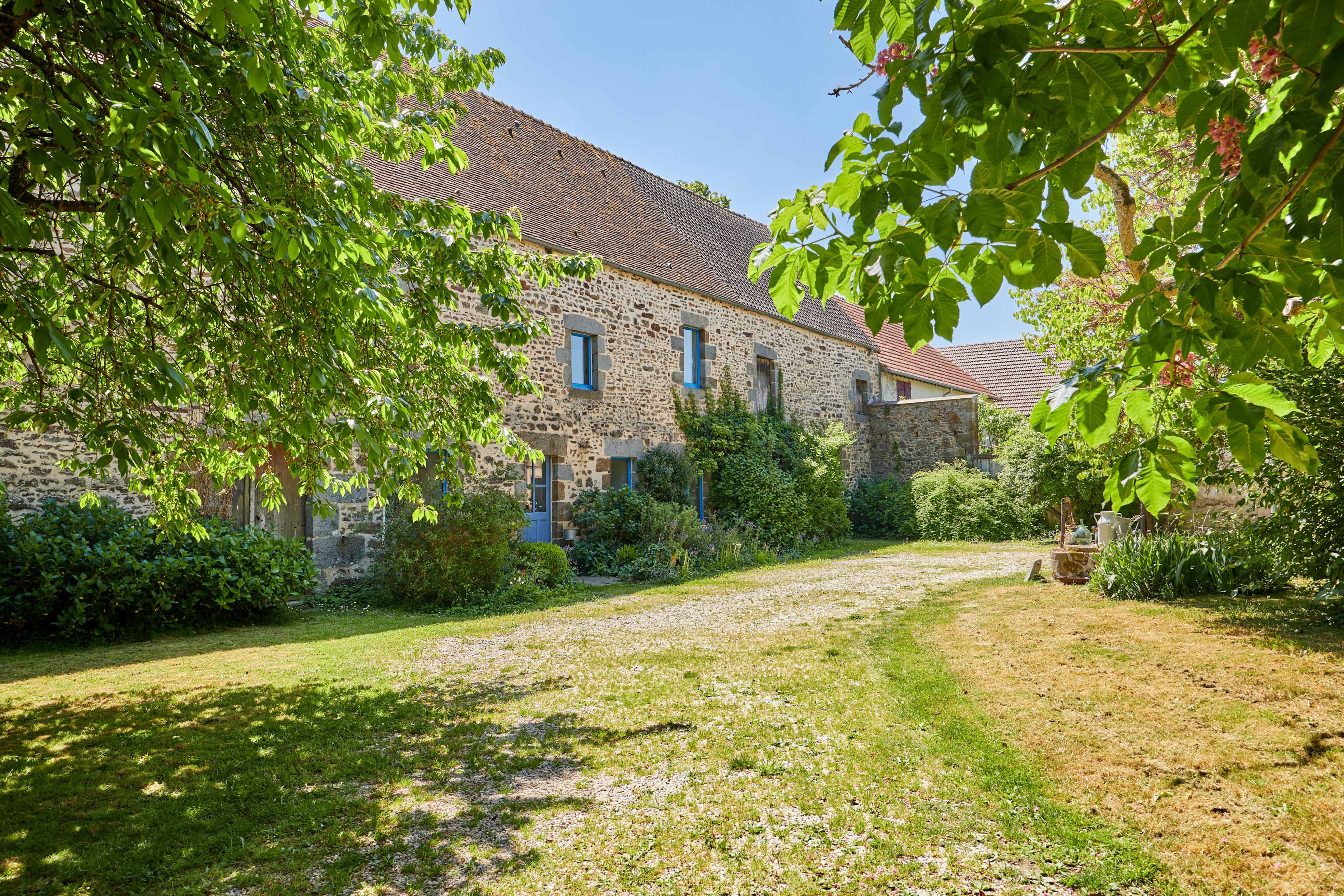 The image shows a stone house surrounded by greenery and a gravel pathway, under a clear blue sky.