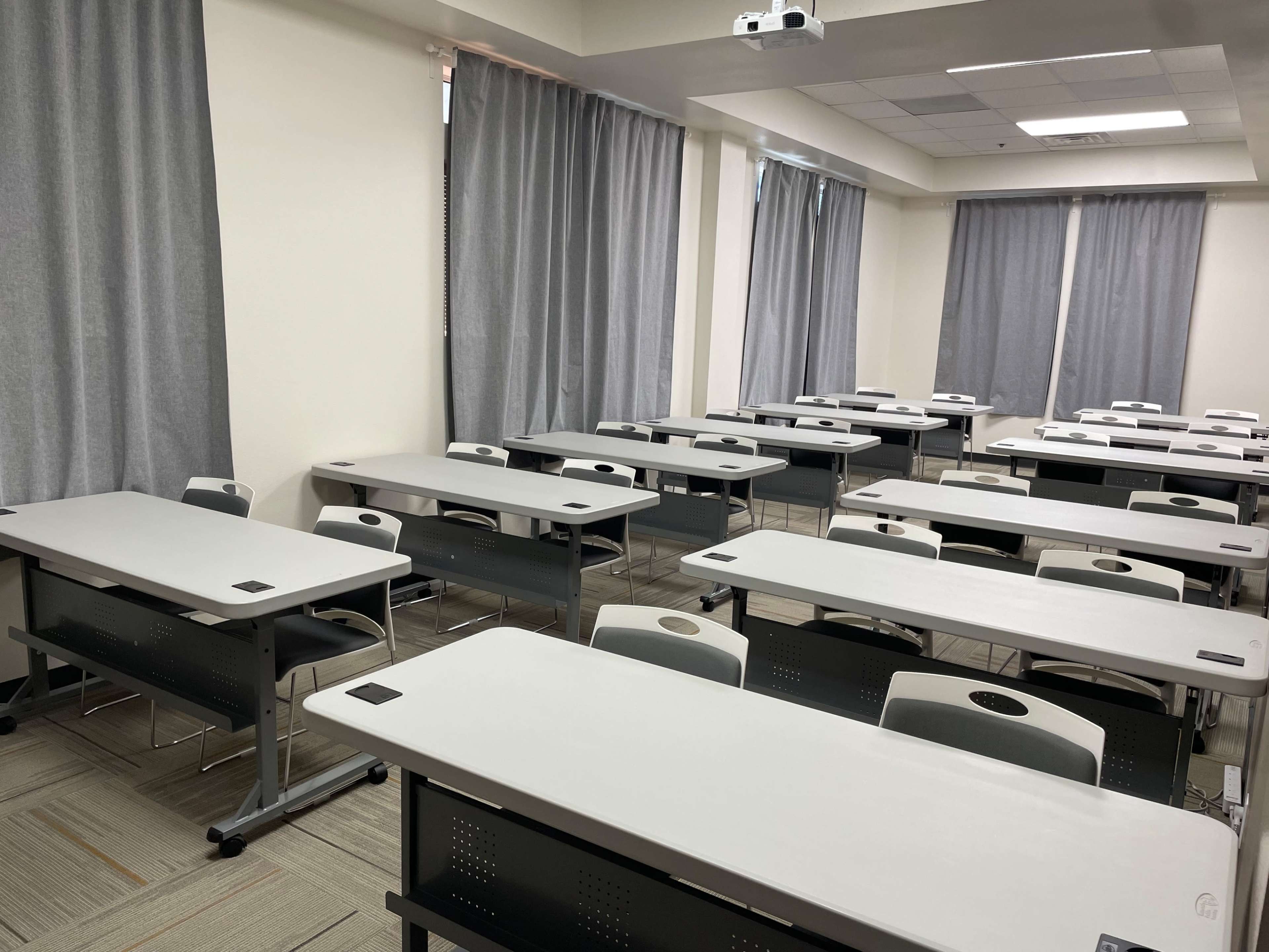 The image shows a classroom with several rows of empty, light-colored desks and gray curtains on the windows.