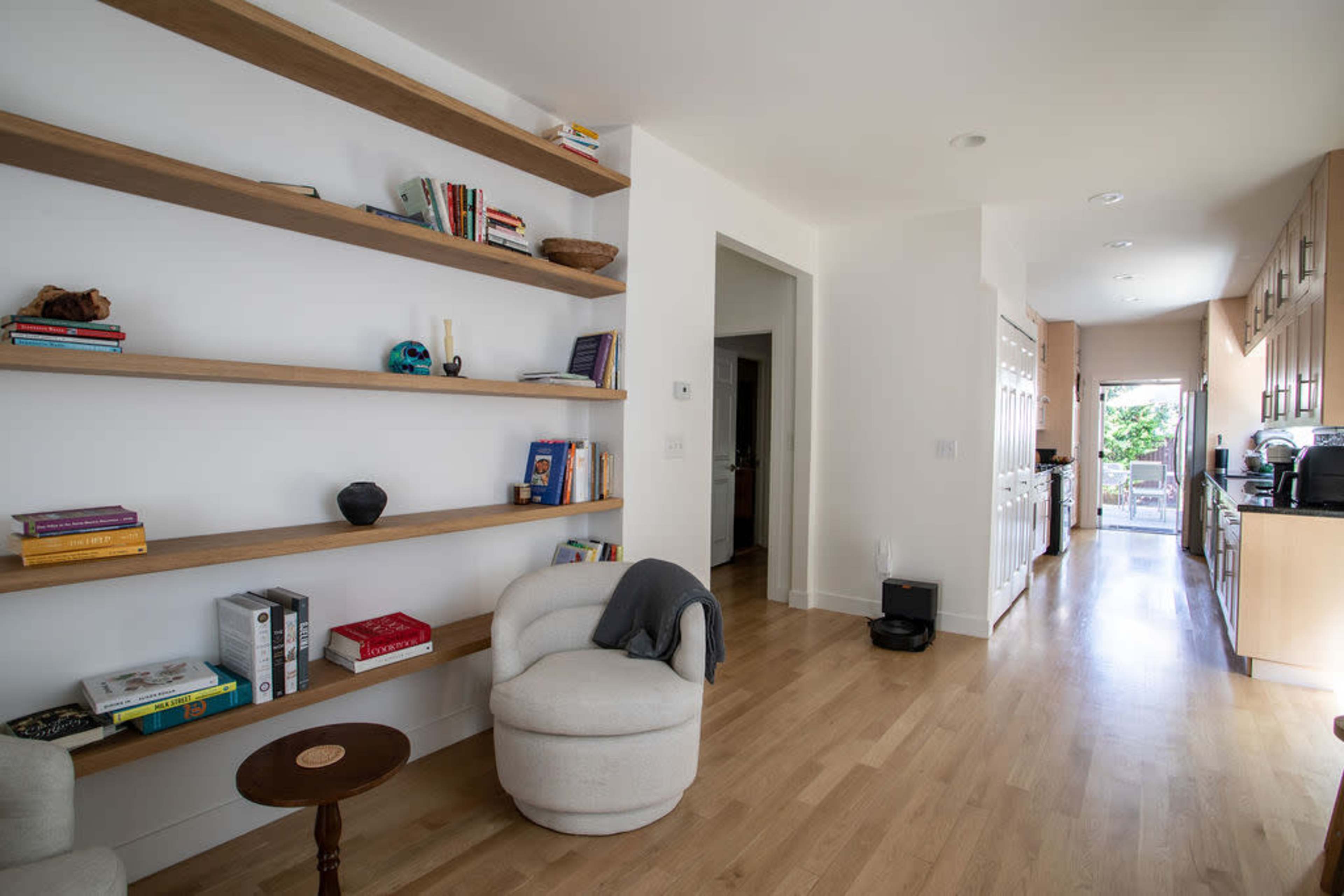 A brightly lit room features a wooden shelf stacked with books and decorative items, alongside a small table and two plush chairs, leading to a hallway that opens into a kitchen area.