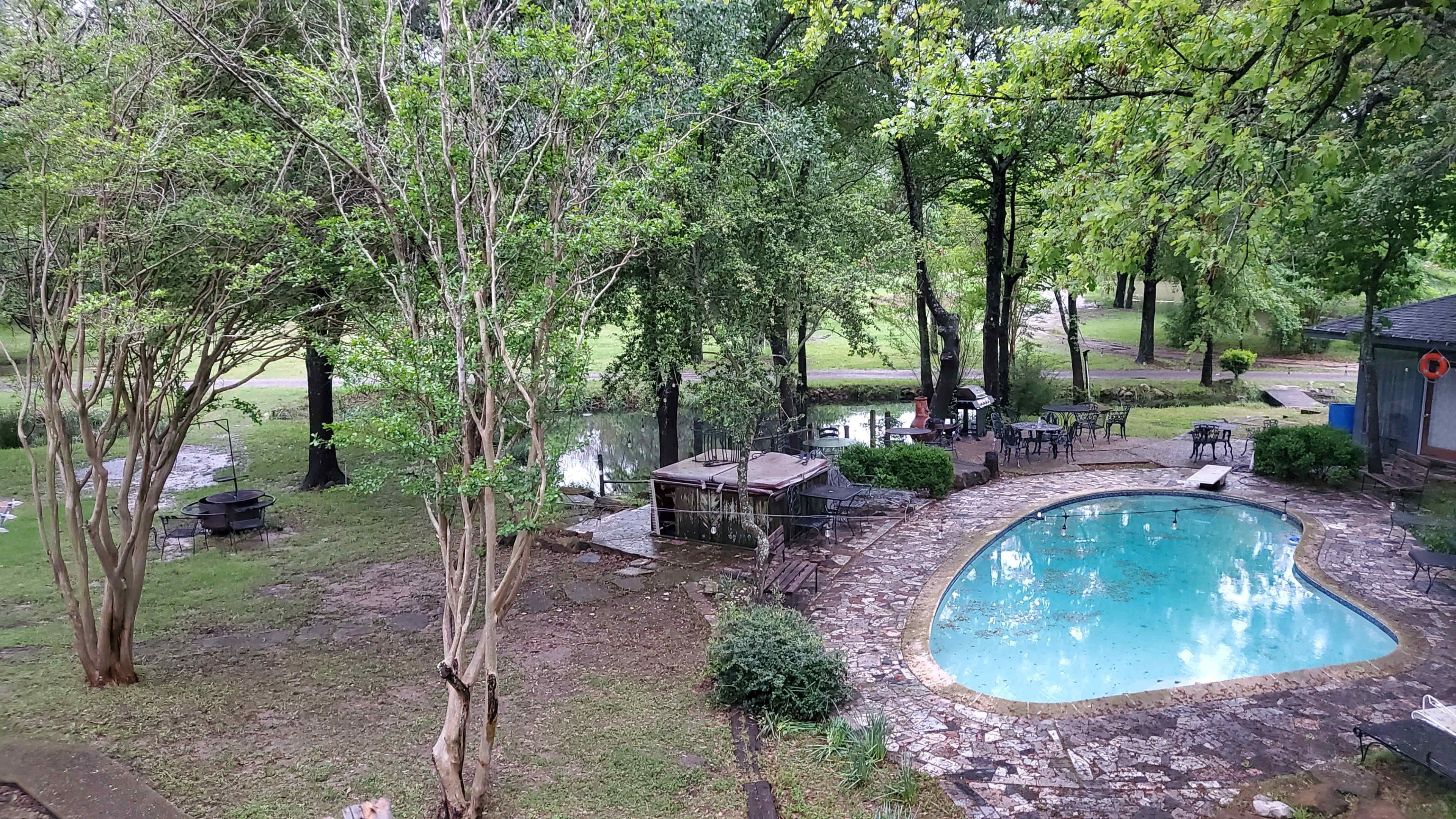 A backyard with a swimming pool, a gazebo, and a grassy area alongside a small body of water, surrounded by trees.