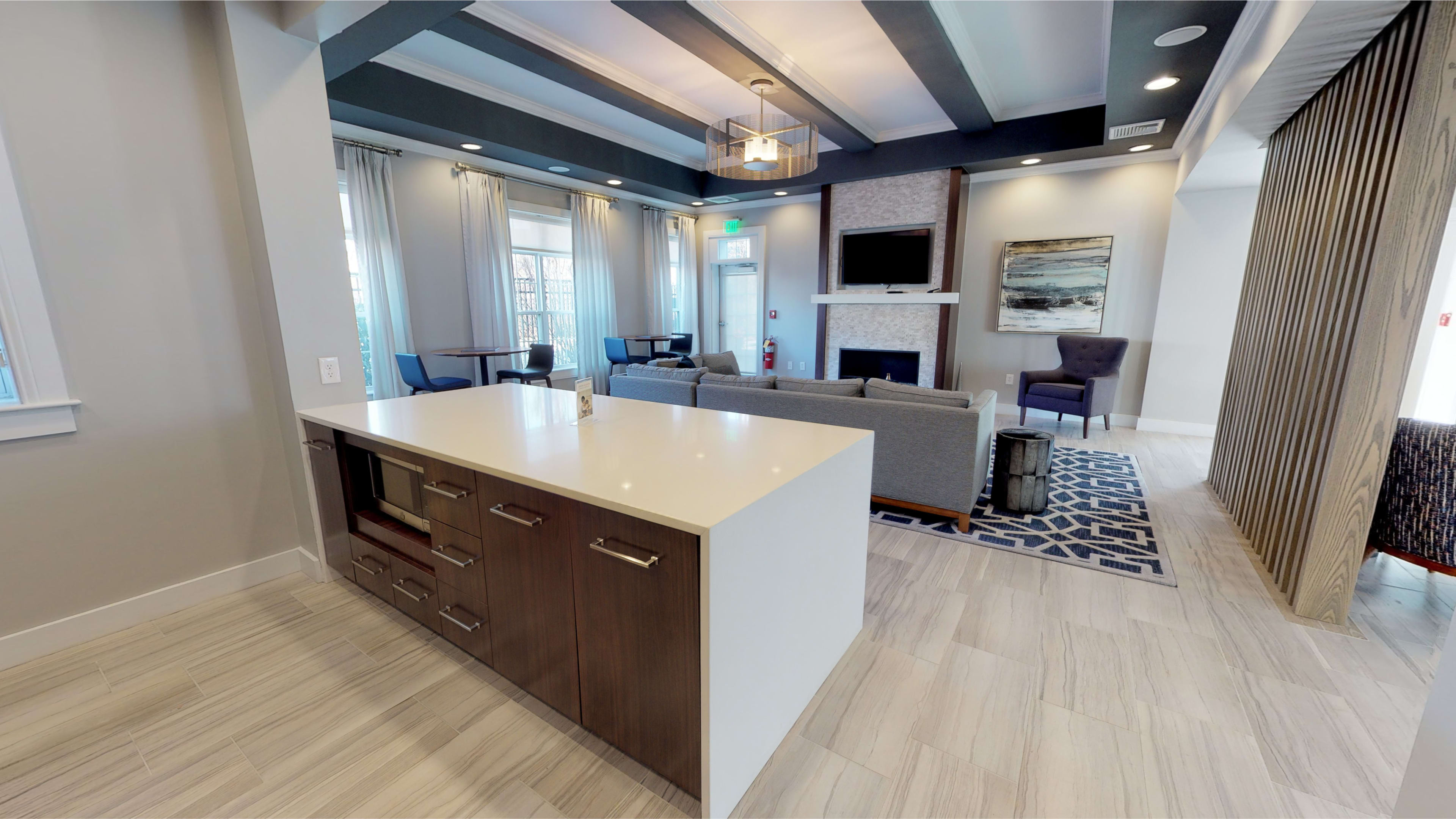 A modern lobby area featuring a kitchen island with cabinetry, seating arrangements, a fireplace, and large windows allowing natural light.
