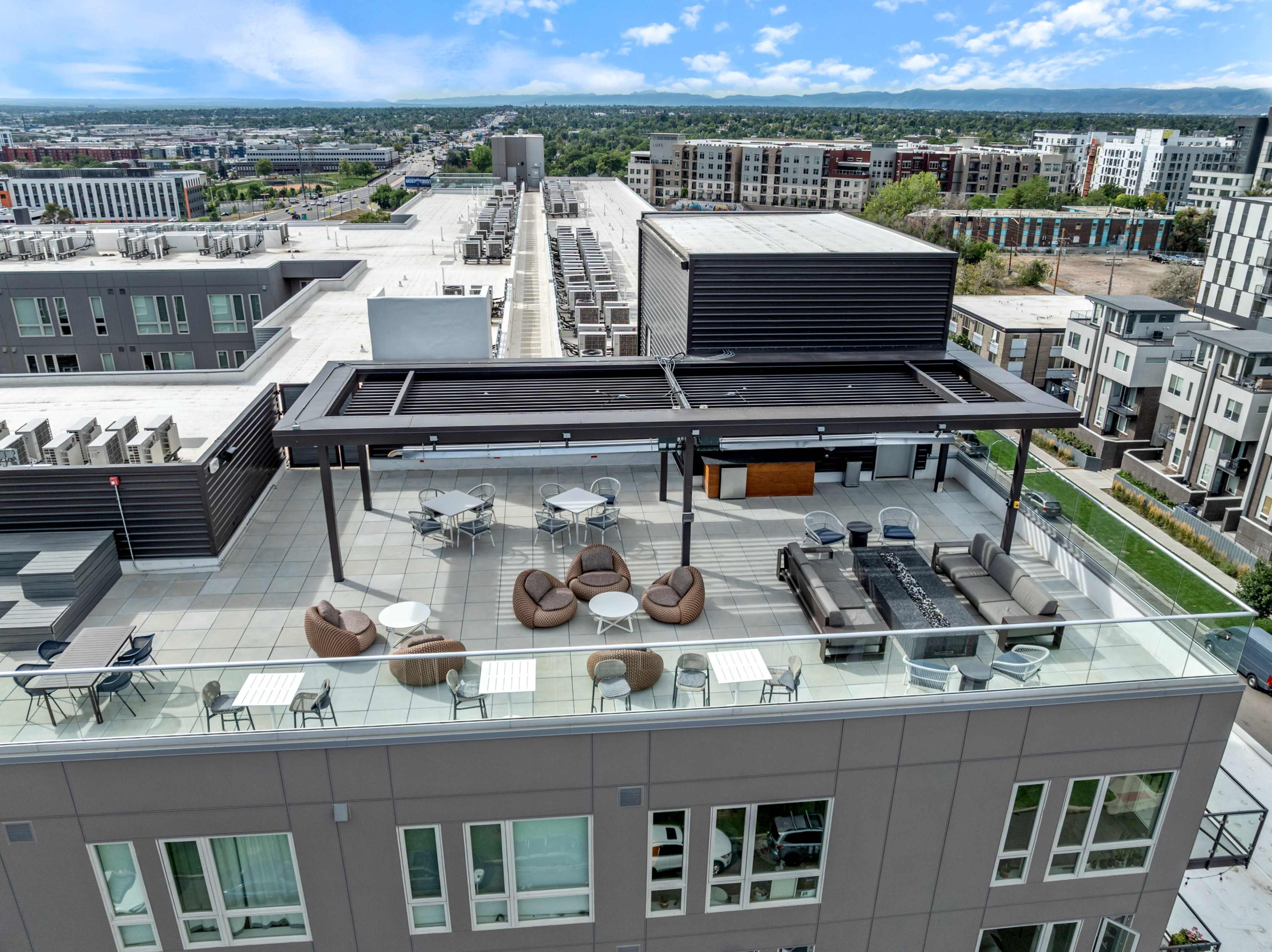 The image shows a rooftop terrace with seating areas, tables, and a fire pit, overlooking a city landscape.