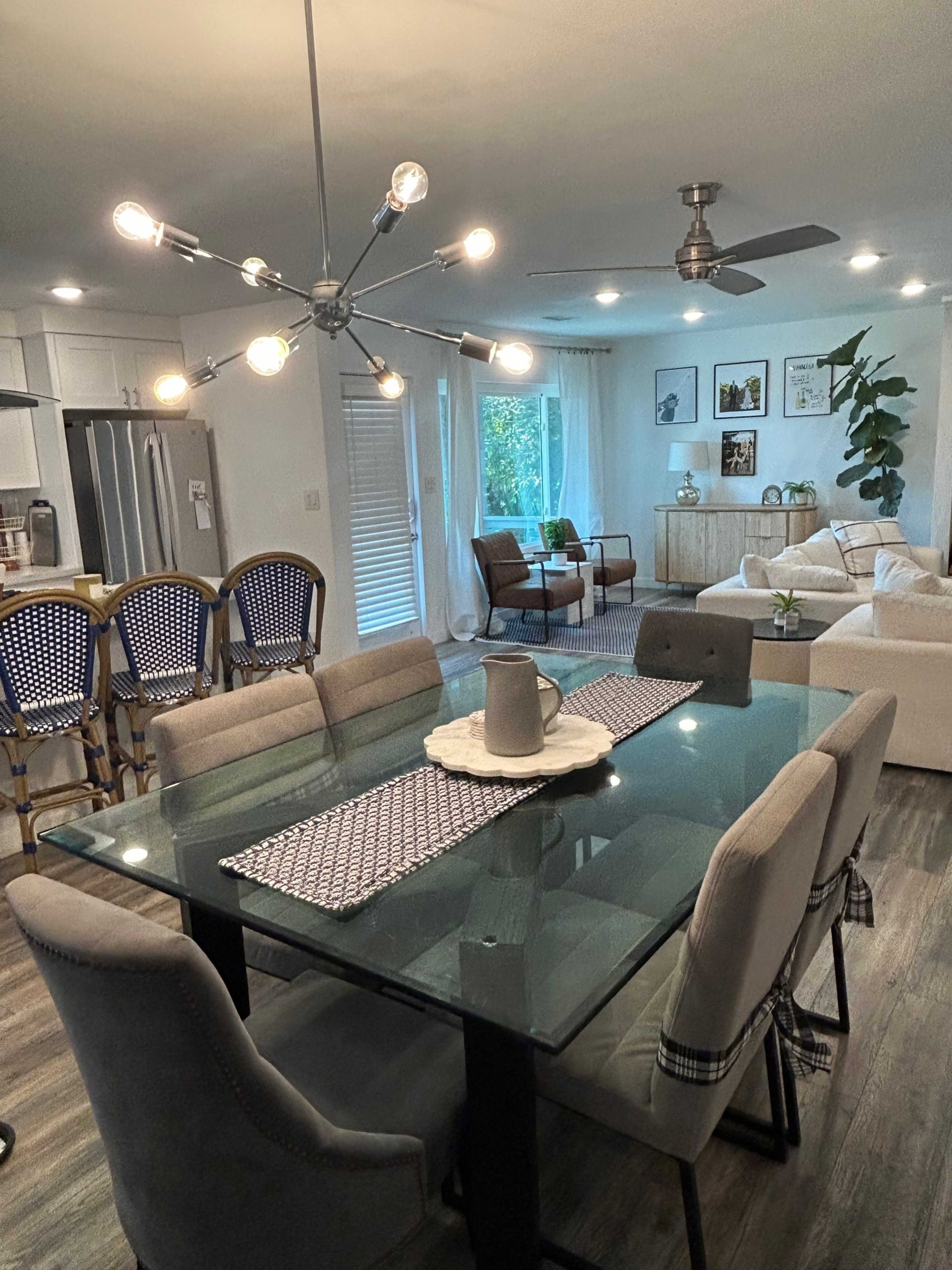 The image shows a modern dining area with a glass table surrounded by upholstered chairs, connected to a kitchen featuring stainless steel appliances and a living room with a white sofa and greenery.