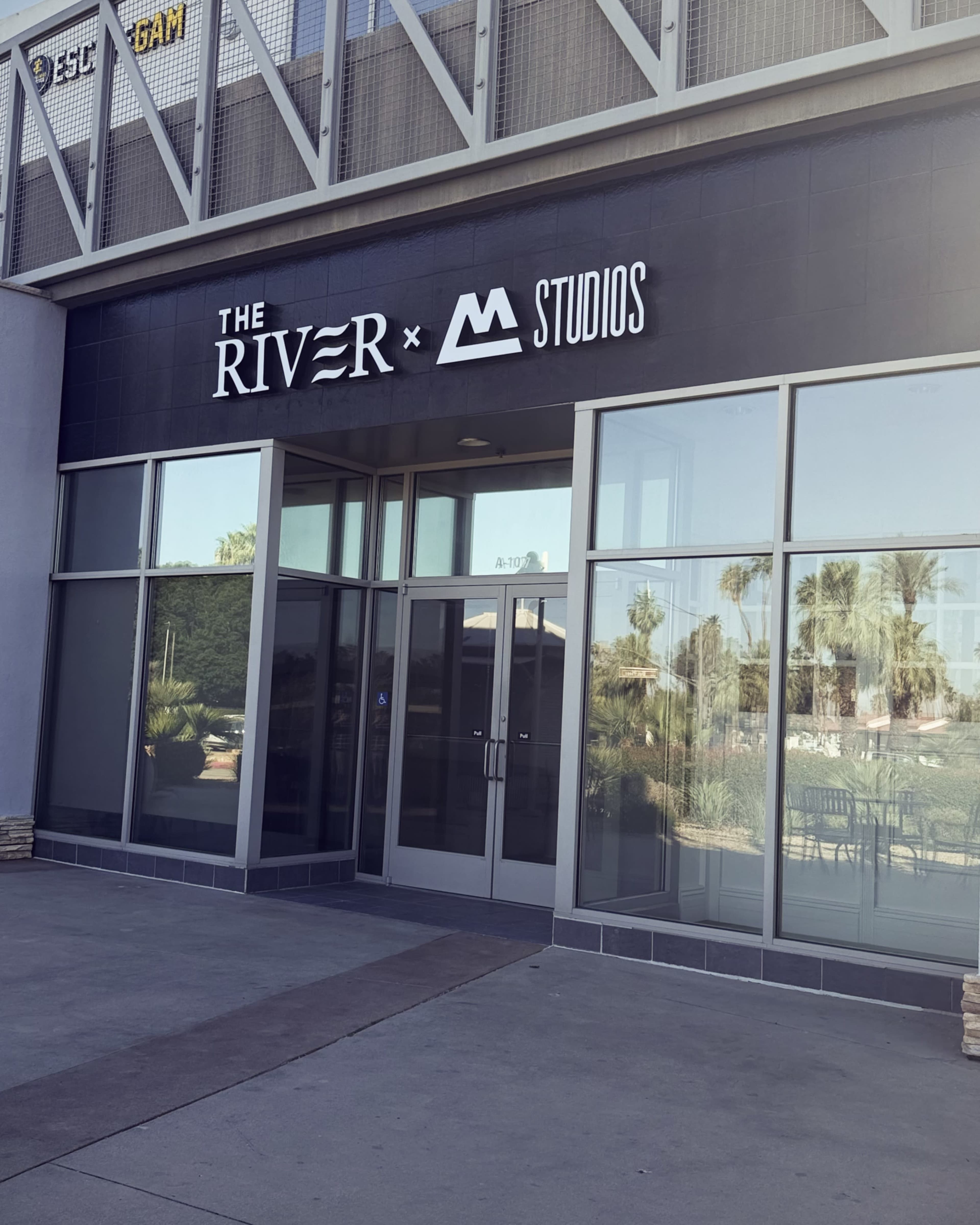 The image shows the entrance of a modern building featuring a sign that reads "THE RIVER x M STUDIOS."